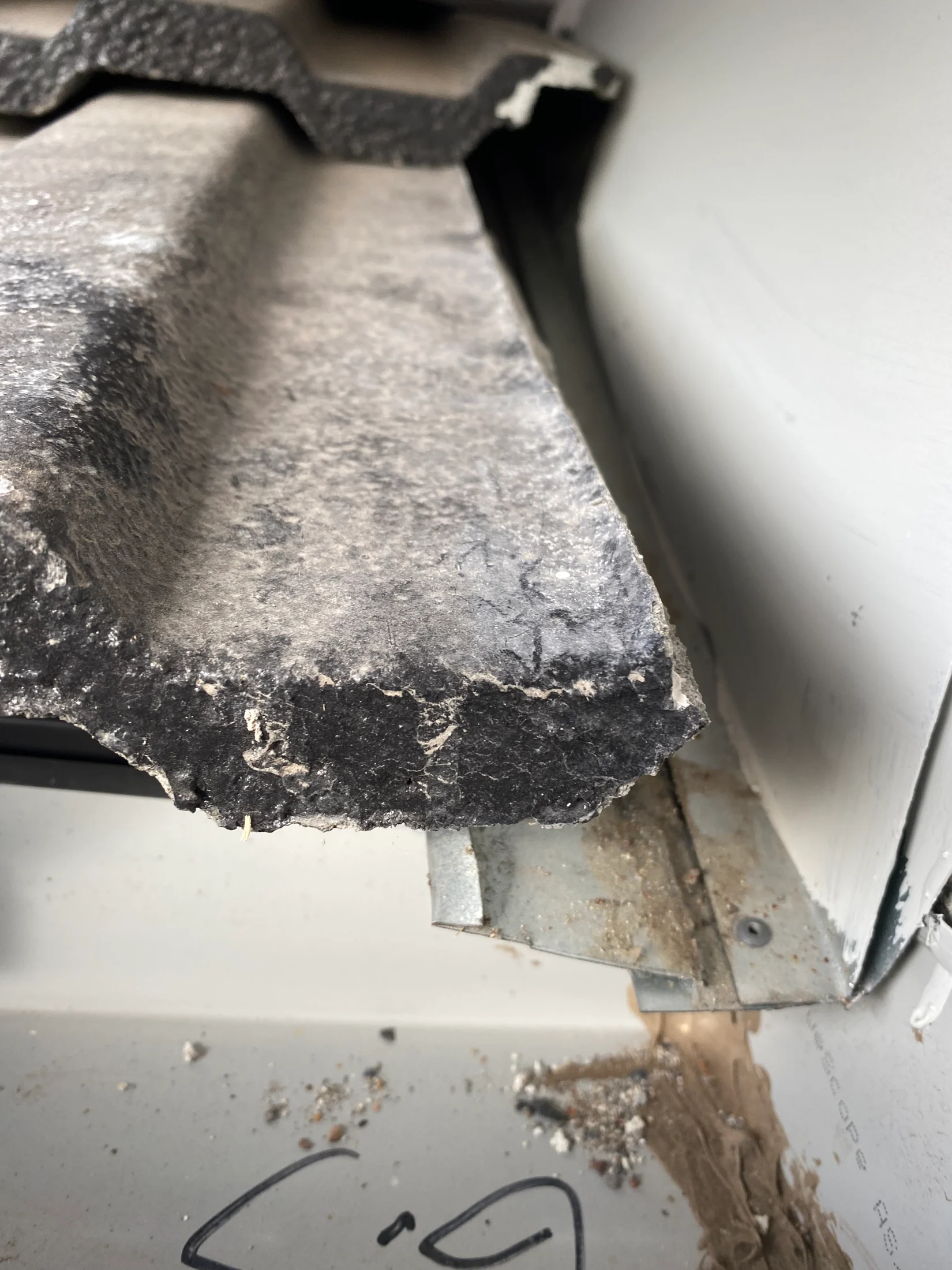 Close-up of a damaged, gray cement roof tile with visible debris and dirt. A metal bracket is partially visible. Edges appear worn.