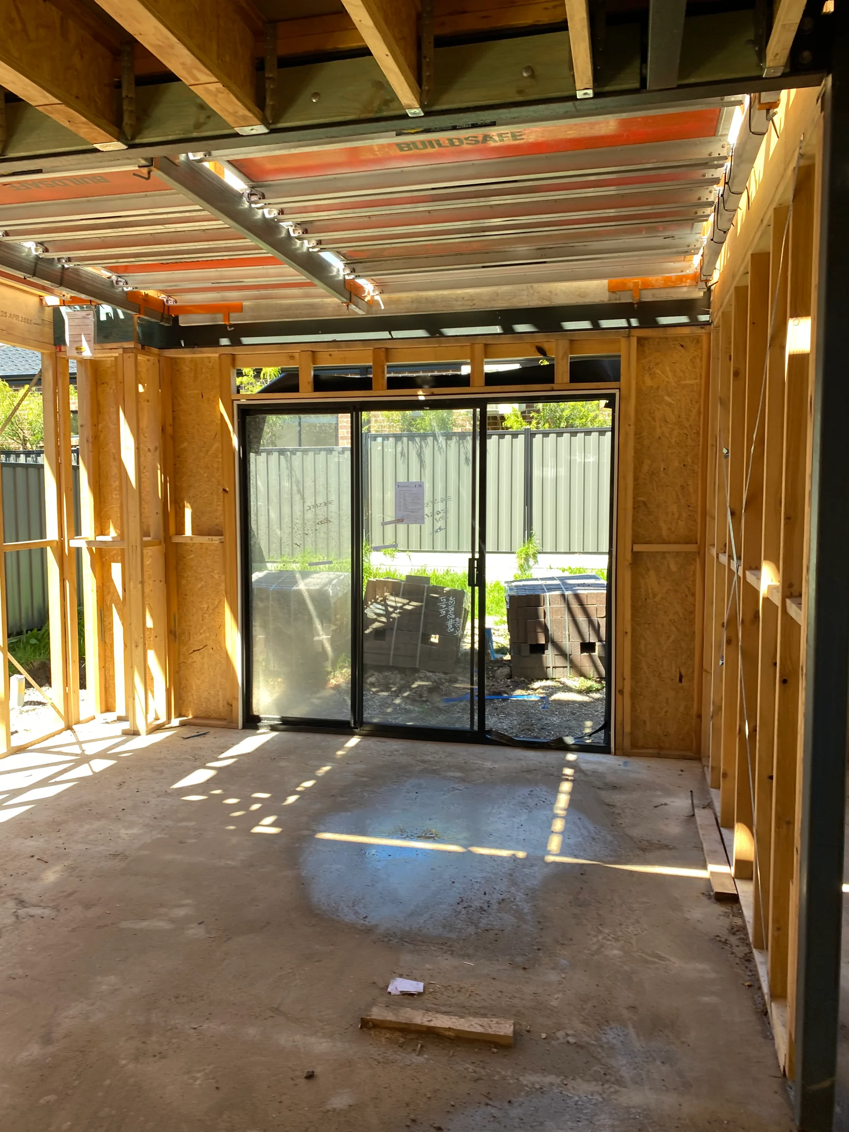 Construction site interior with exposed wooden beams, sliding glass doors, and sunlight casting shadows on the concrete floor.