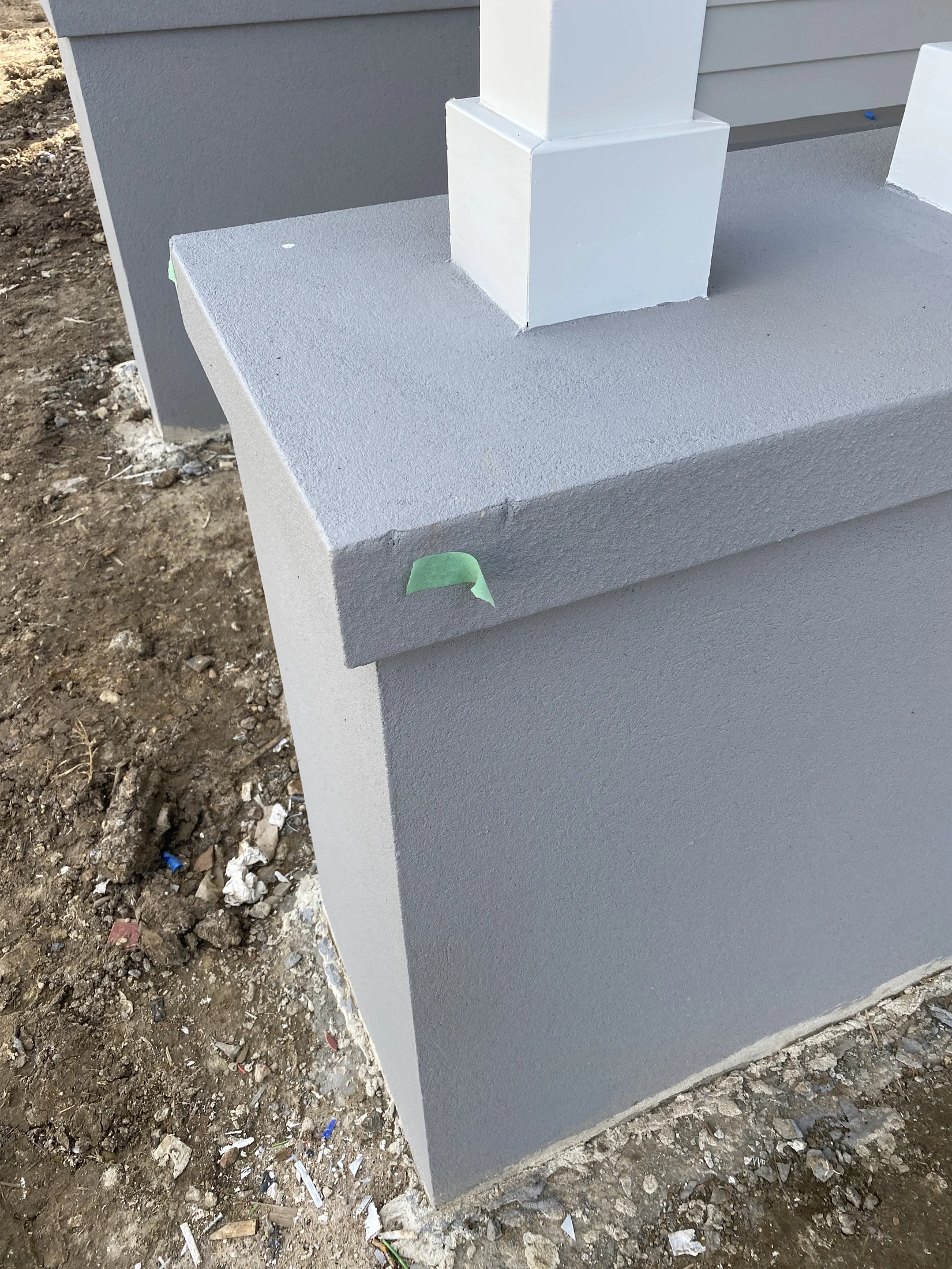 Gray concrete pillar with white top, green tape on edges, set on a dirt ground with scattered debris. Overcast lighting.