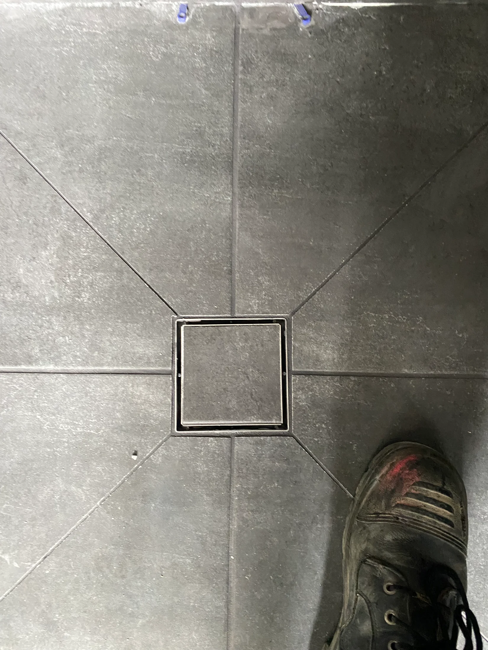 A black boot stands on dark gray tiled floor with a square drain cover in the center. The floor has light wear marks. Mood is industrial.