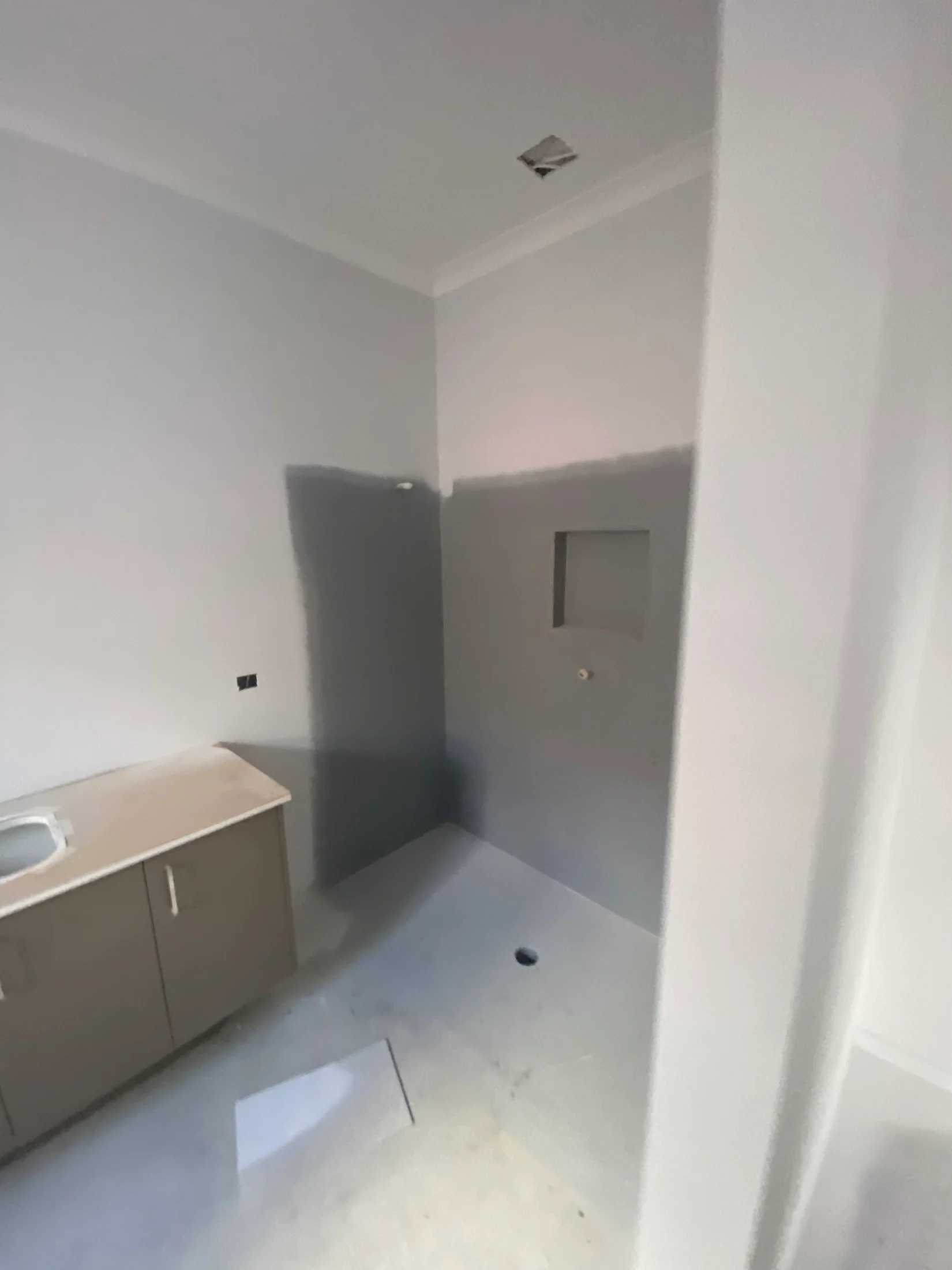 Unfinished bathroom with gray walls, missing sink, light fixture opening. Beige cabinet and a wall niche visible; bare floor and muted tones.