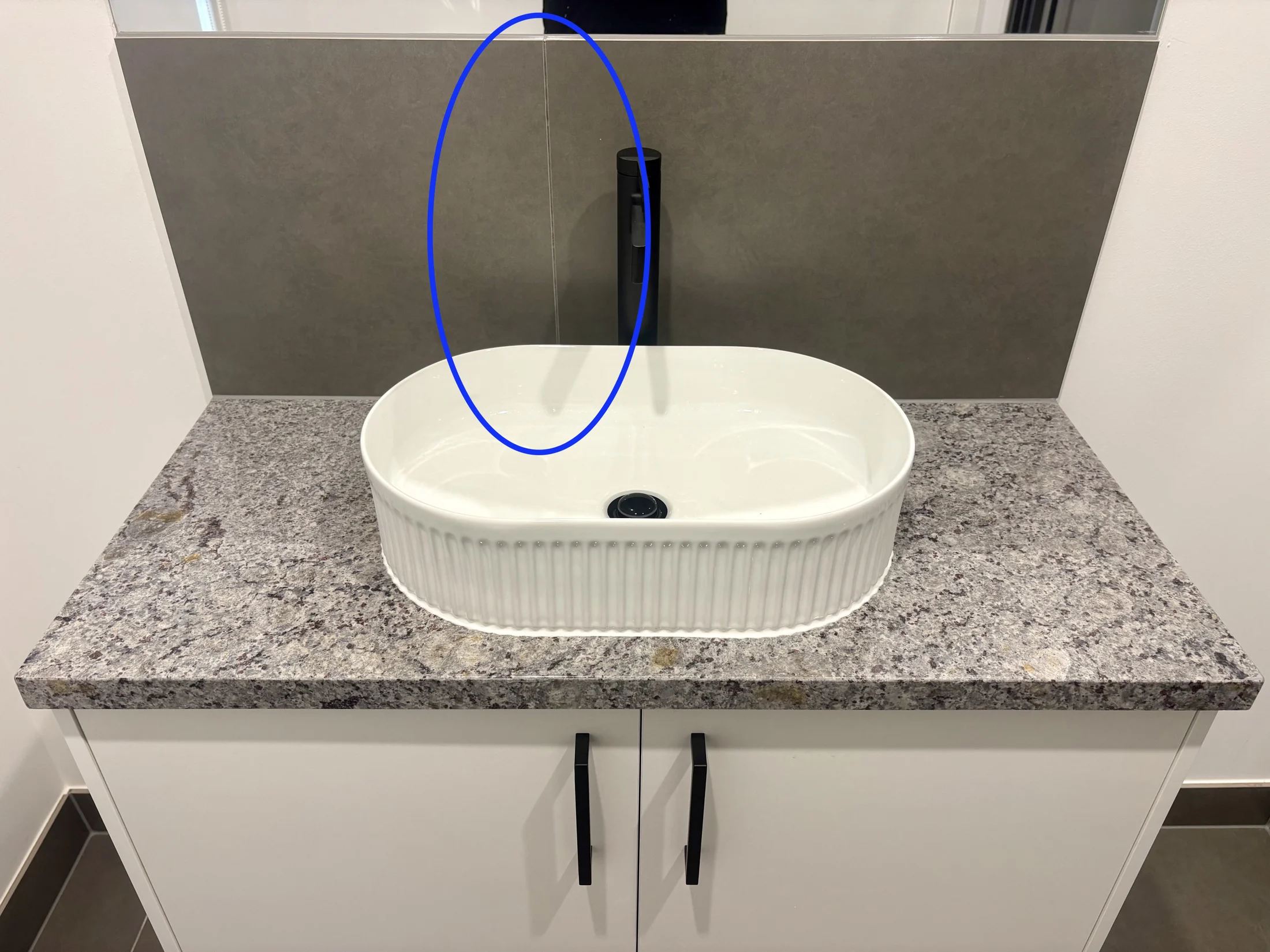 Granite countertop with a white oval sink, black faucet, and gray backsplash. Blue circle highlights area above the sink. Neutral tones.