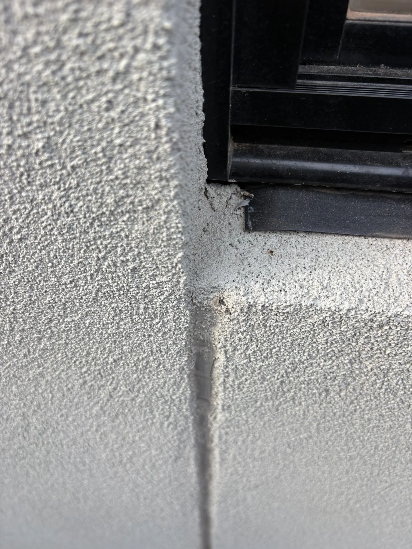 Close-up of a textured wall corner with a small crack near a black window frame. The surface is light gray and slightly rough.