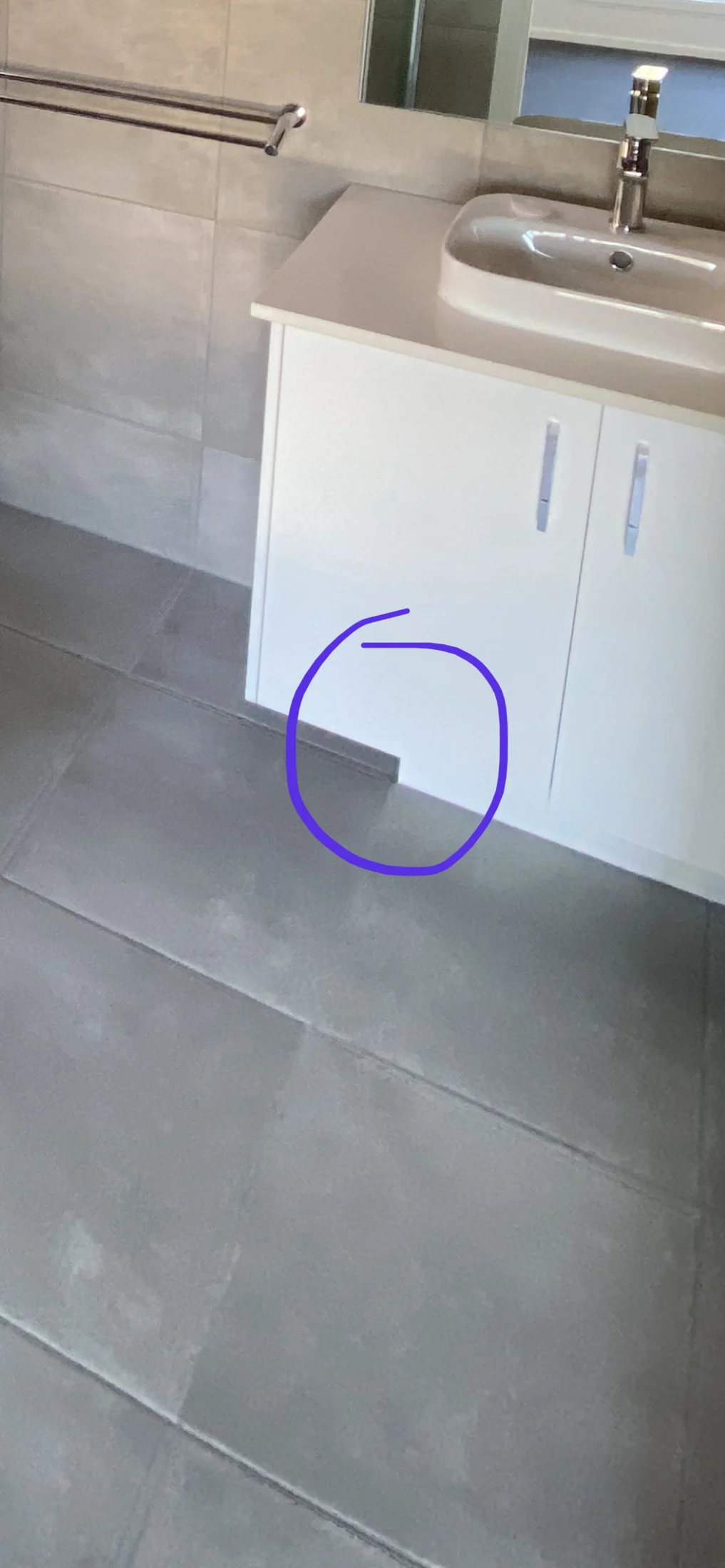 A bathroom scene with gray tiles and a white cabinet under a sink. A purple circle highlights a gap at the bottom edge of the cabinet.