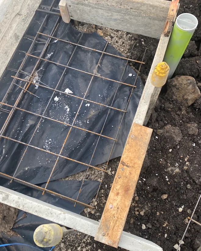 Wooden frame with metal rebar grid set on a black tarp, surrounded by soil. Yellow and green plastic caps on pipes are visible.