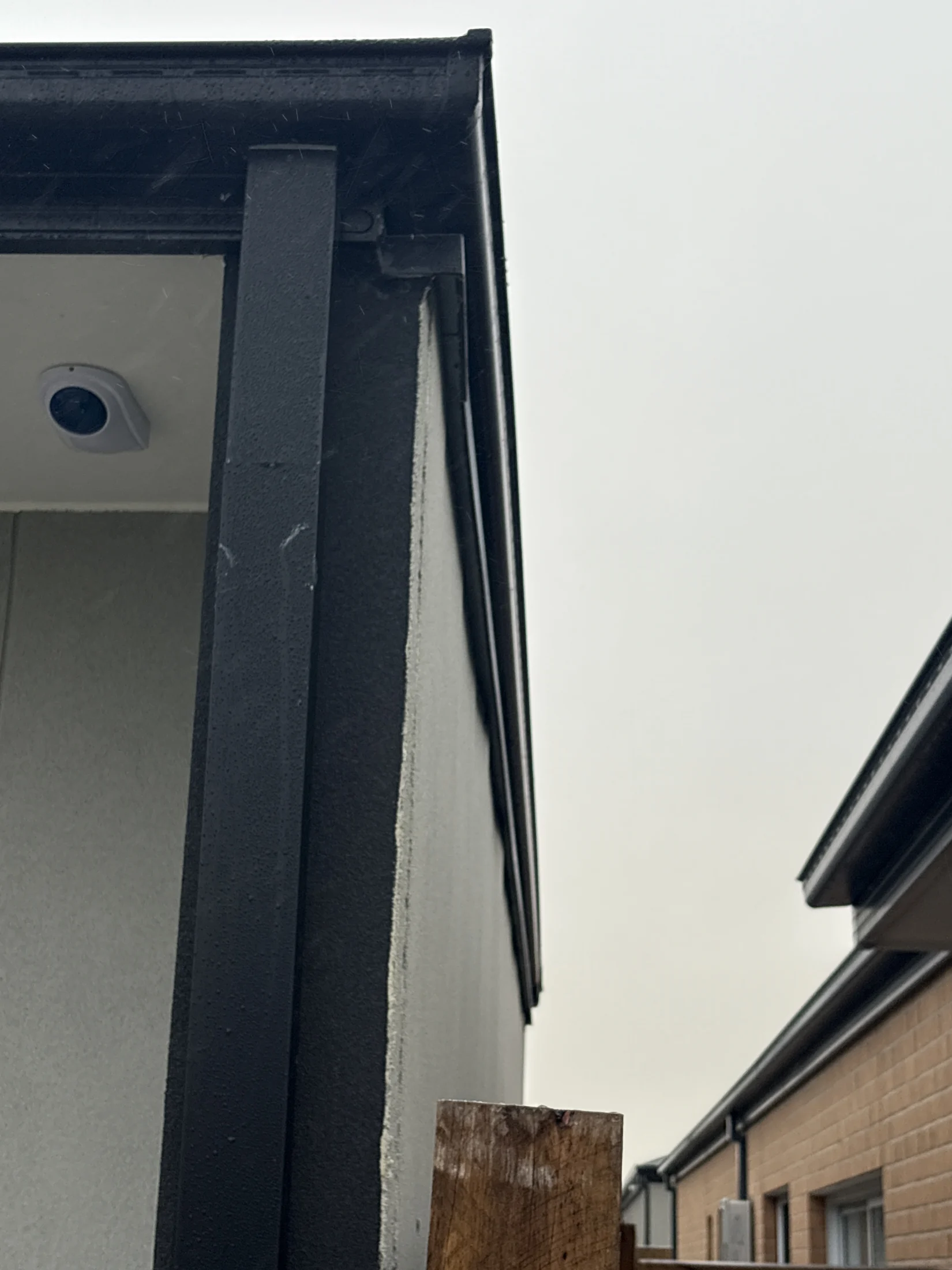 Building corner with black trim, surveillance camera, and adjacent brick wall under overcast sky. Wooden post visible in foreground.
