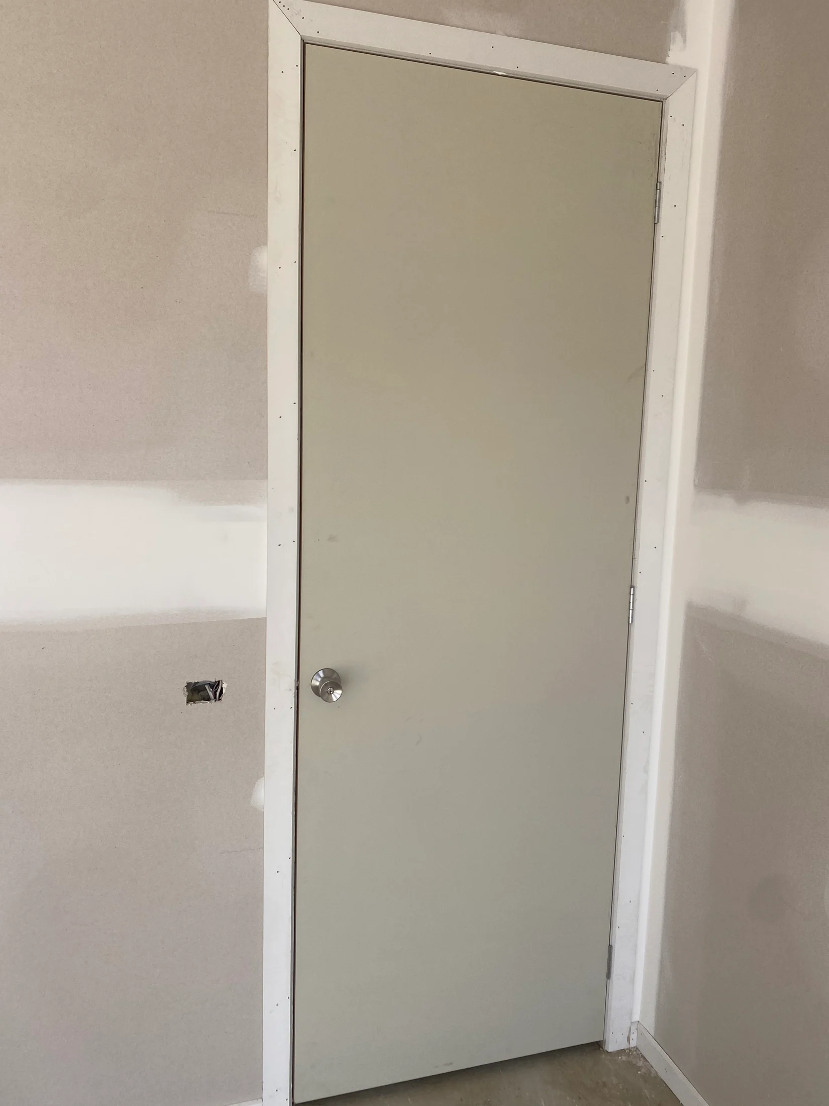 Plain beige door with silver handle in a partially painted room. Walls show drywall texture, with an uncovered electrical outlet. Calm setting.