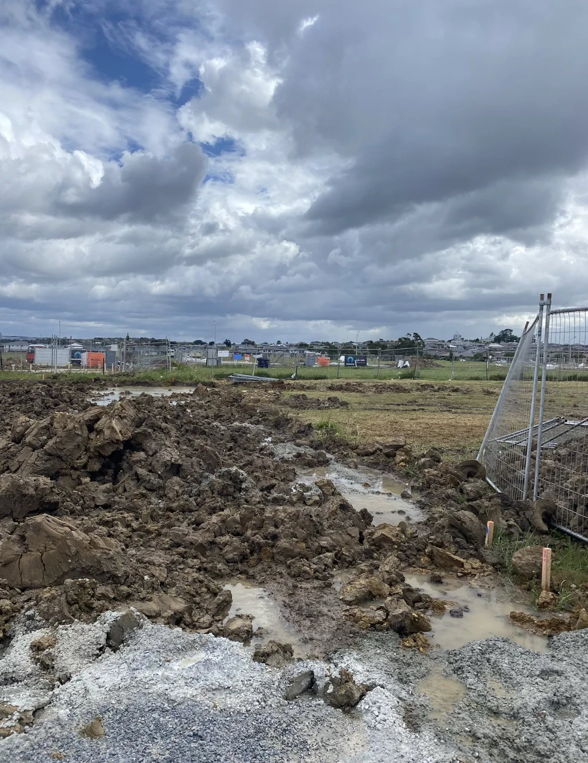 Muddy construction site with puddles, surrounded by a metal fence under a cloudy sky. Distant buildings are visible in the background.