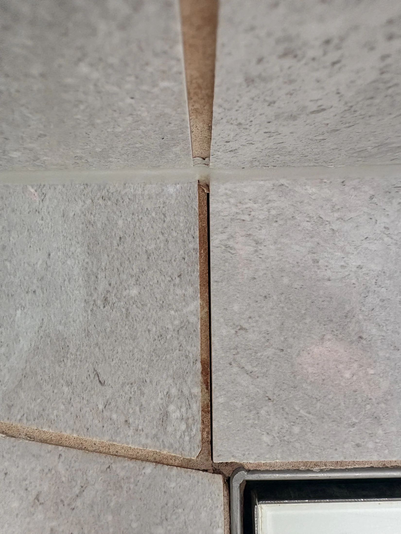 Close-up of gray, textured tiles with visible grout lines. A white object is partially visible in the bottom right corner.