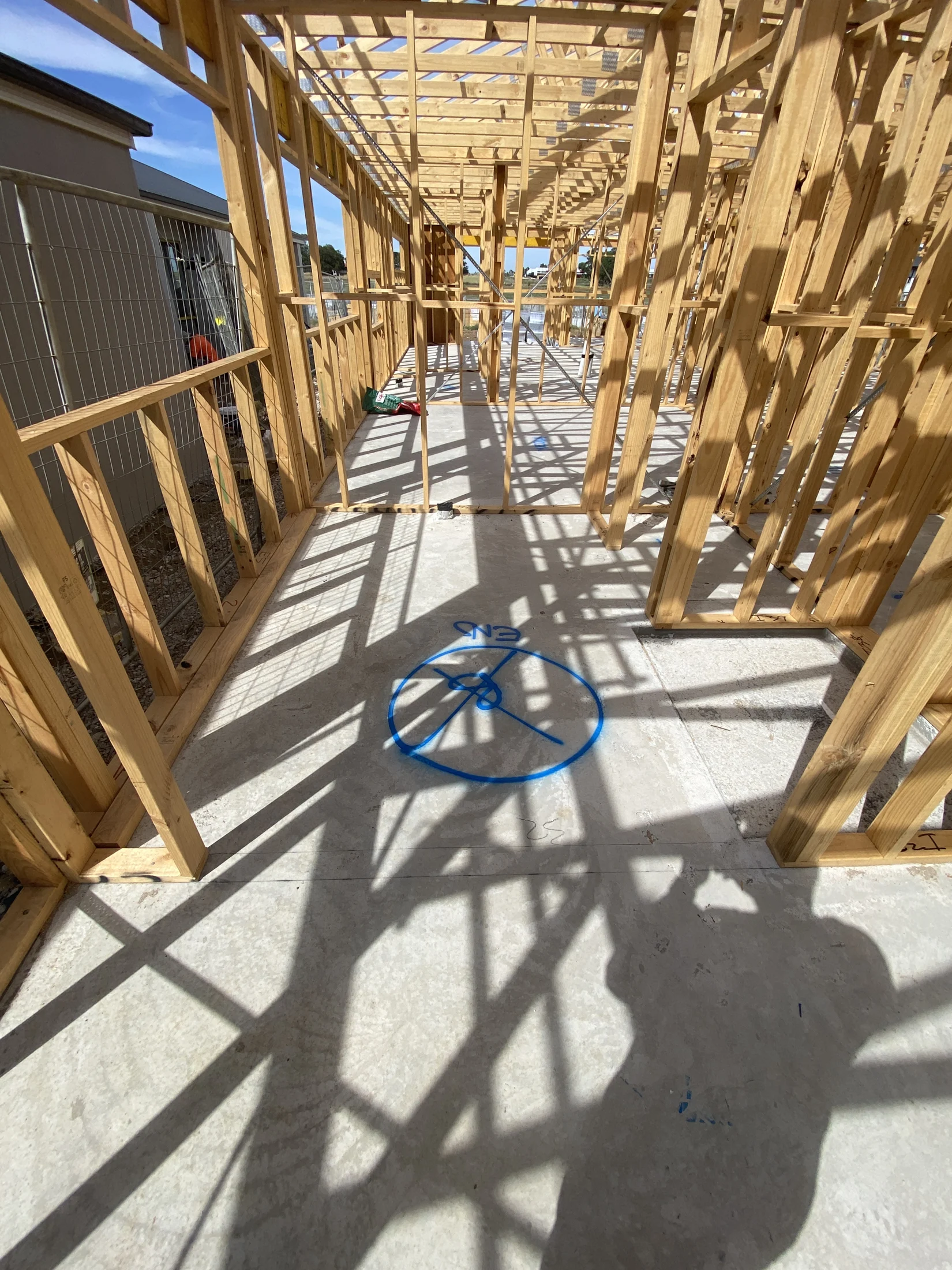 Wooden frame of a house under construction, with blue circle marking on concrete floor, casting shadows in bright sunlight. No text visible.