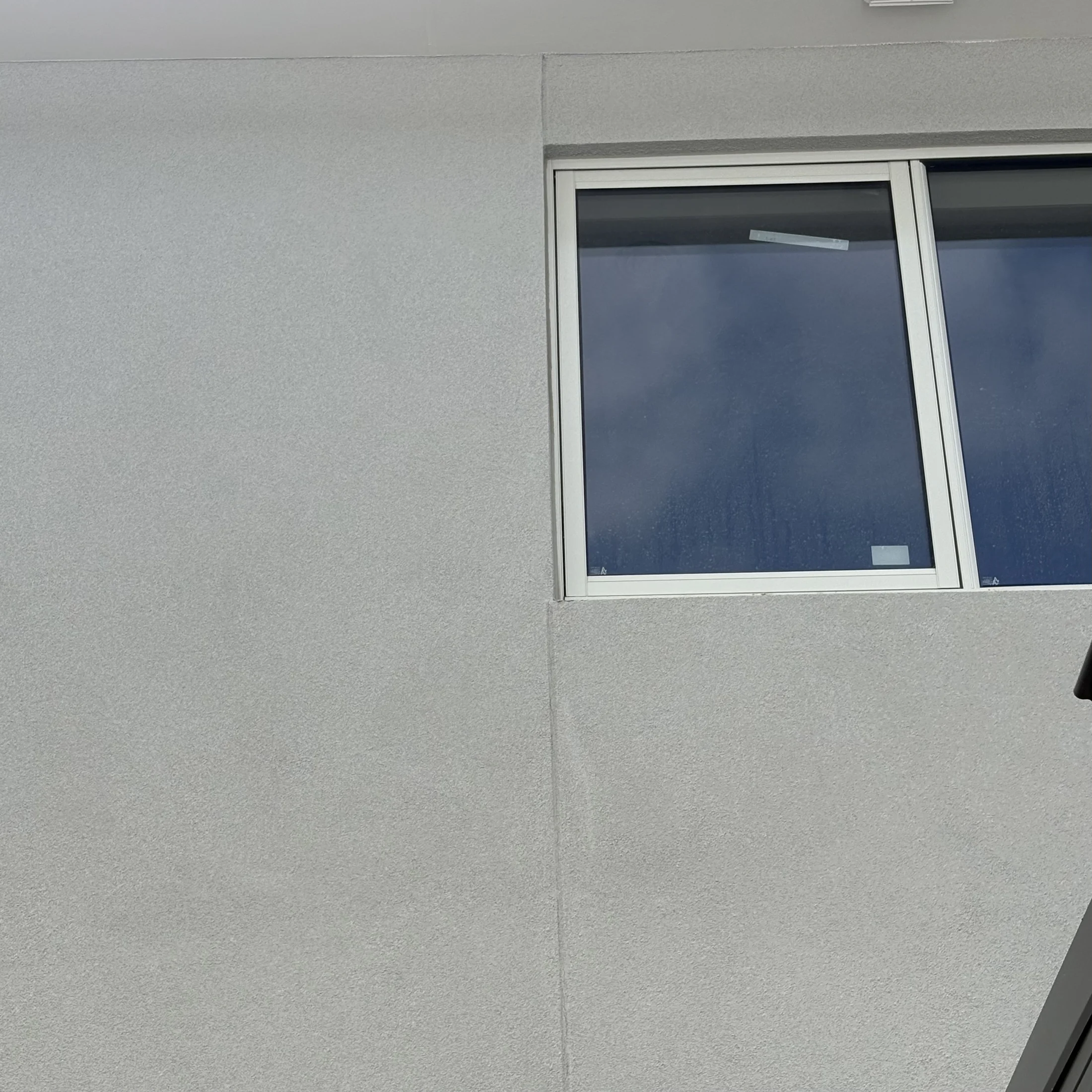 A gray wall with a partially visible window showing a cloudy sky. A small part of a black object is visible in the bottom right corner.