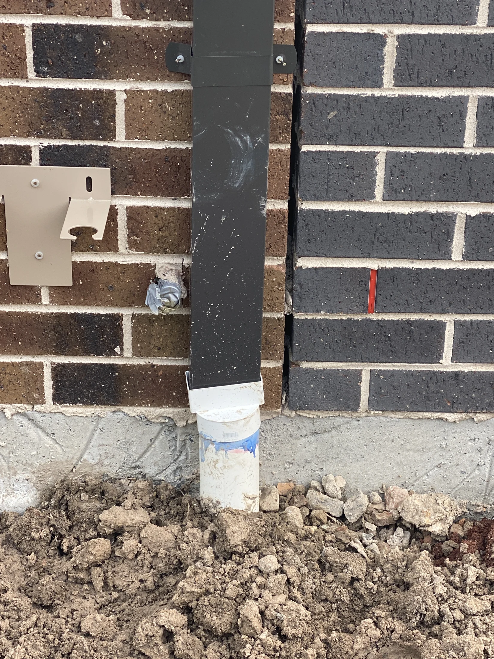 Brick wall with a black downspout and white pipe. Dirt and rocks at the base. Brown bricks on the left, dark grey on the right. No visible text.
