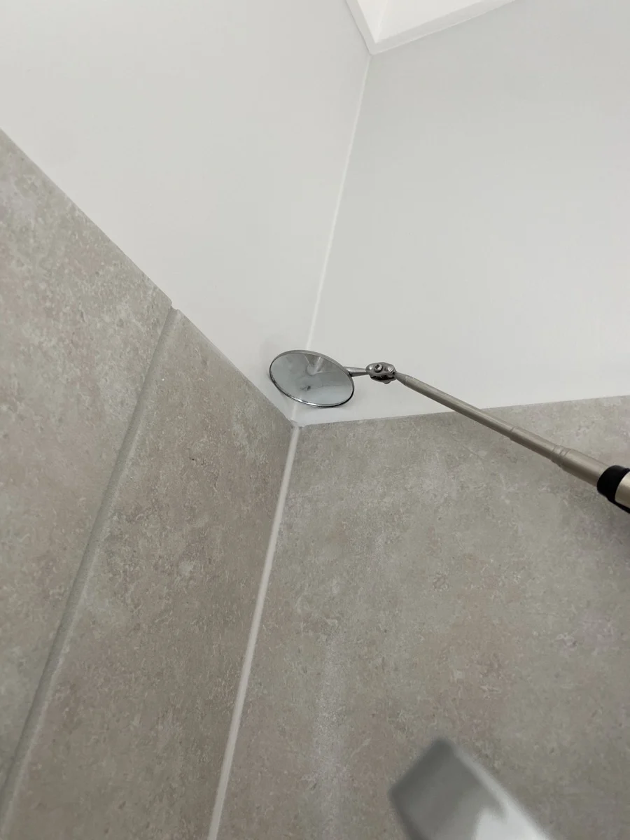 Tile shower corner with a telescopic inspection mirror reflecting the junction. Gray tiles and white ceiling create a clean, minimal look.
