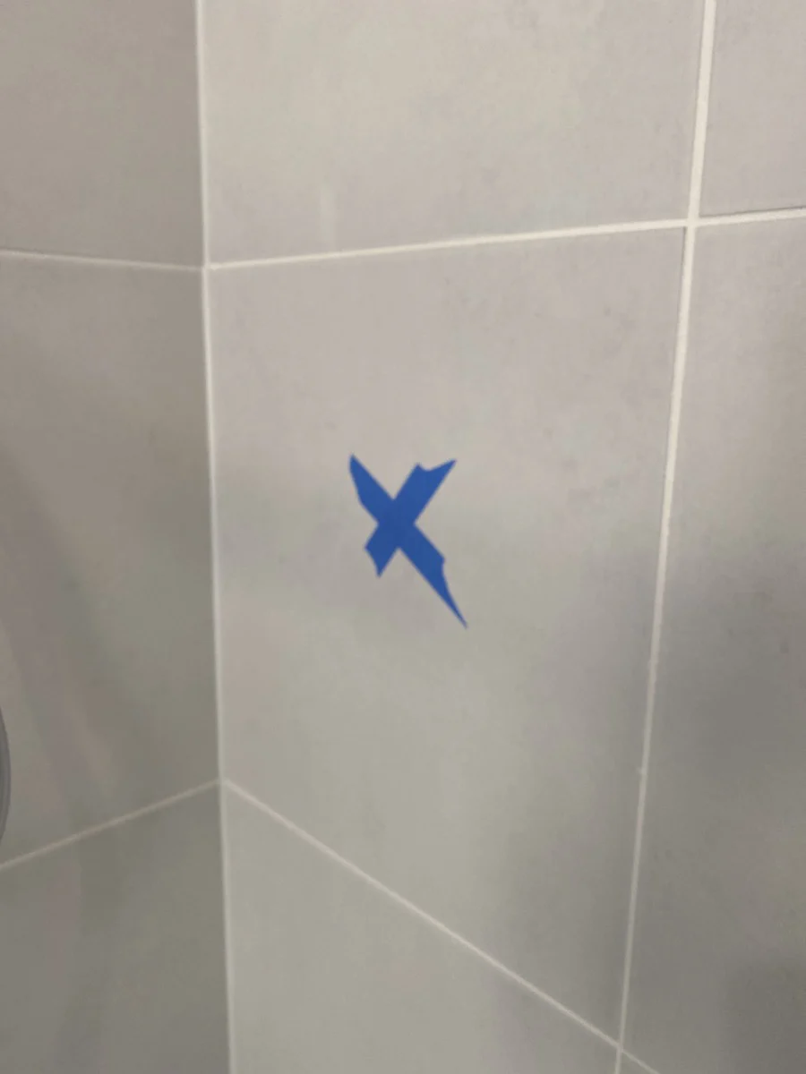 Blue tape in an "X" shape on a tiled white wall. The setting appears neutral, with a focus on the taped marking.