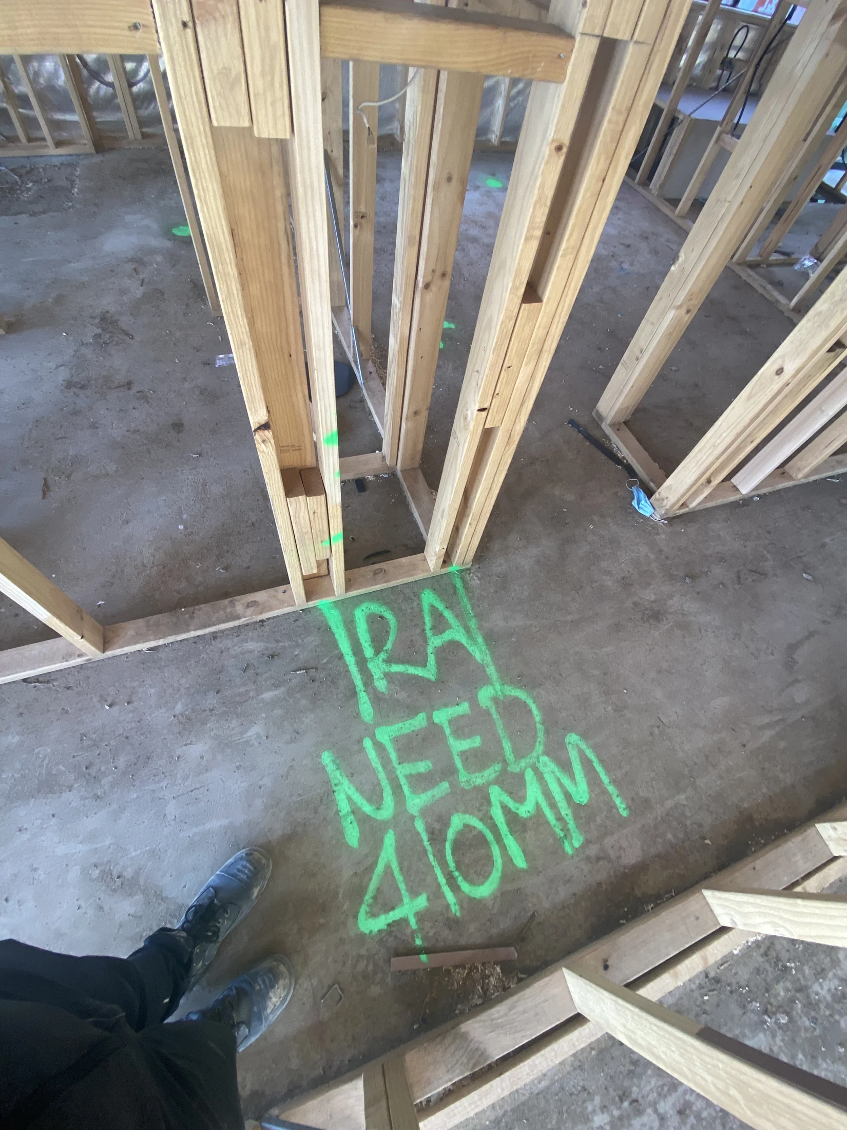 Wooden frame on a construction site with "Ira need 410mm" in bright green paint on the floor. Person in sneakers stands nearby.