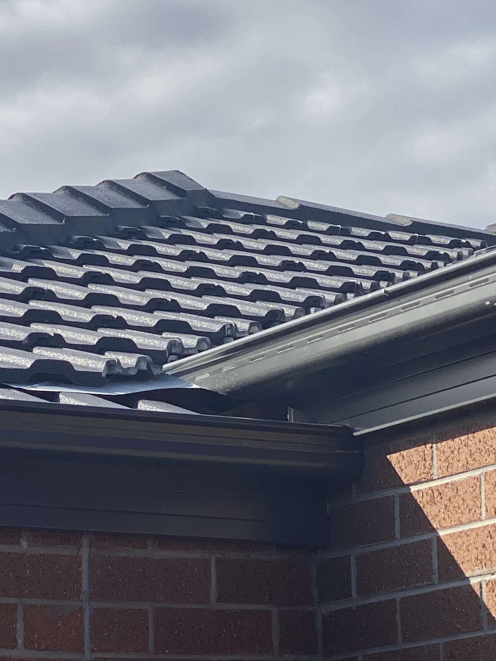 Close-up of a dark gray metal roof with brick wall under cloudy sky. The sun casts shadows, highlighting the roof's wavy texture.