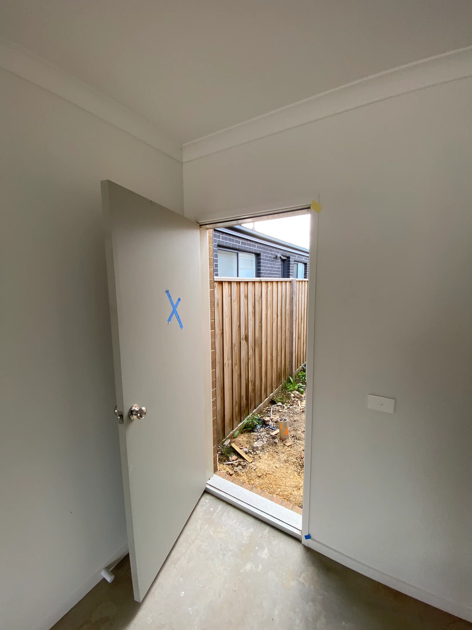 Open door with a blue "X" in a white room leading to a backyard. Wooden fence and adjacent house in view, with earthy ground outside.
