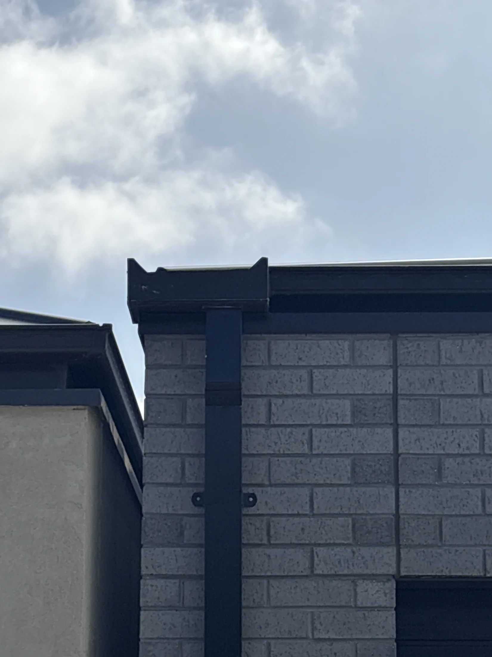 Building corner with grey brick and cream wall, set against a cloudy blue sky. Dark gutter and downspout on the edge. Calm mood.