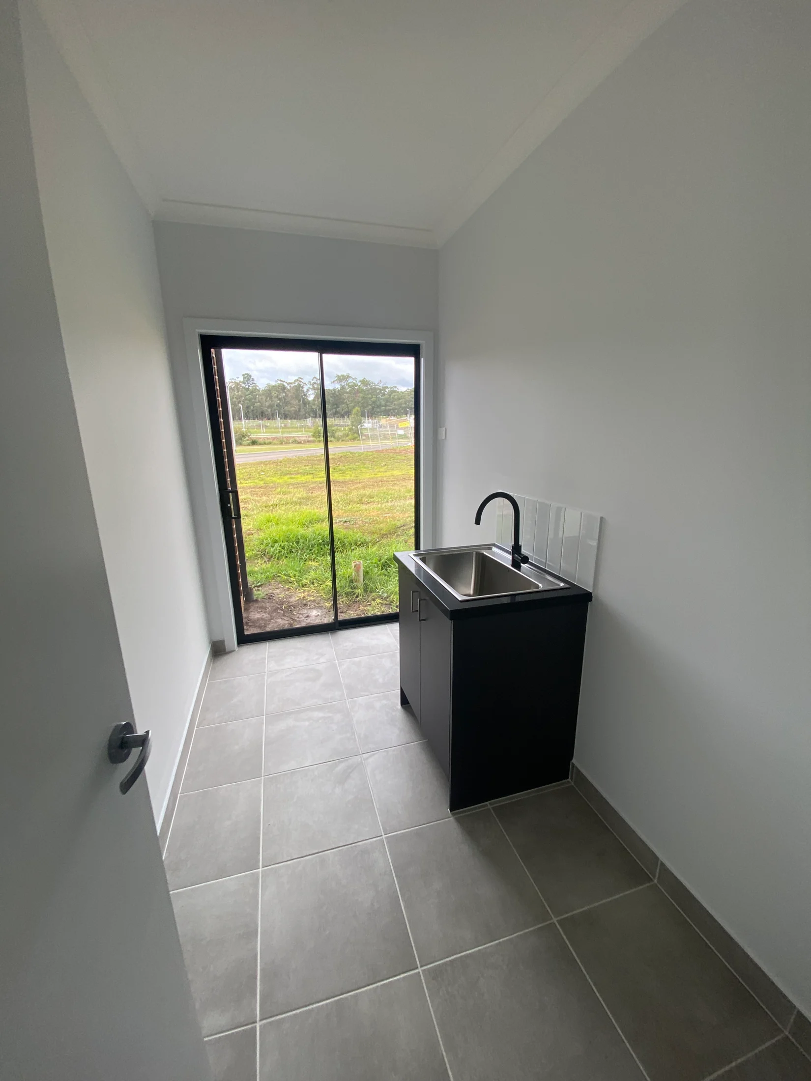 Laundry room with a dark sink, gray tiled floor, and large window showing green grass outside. Bright and minimalistic space.