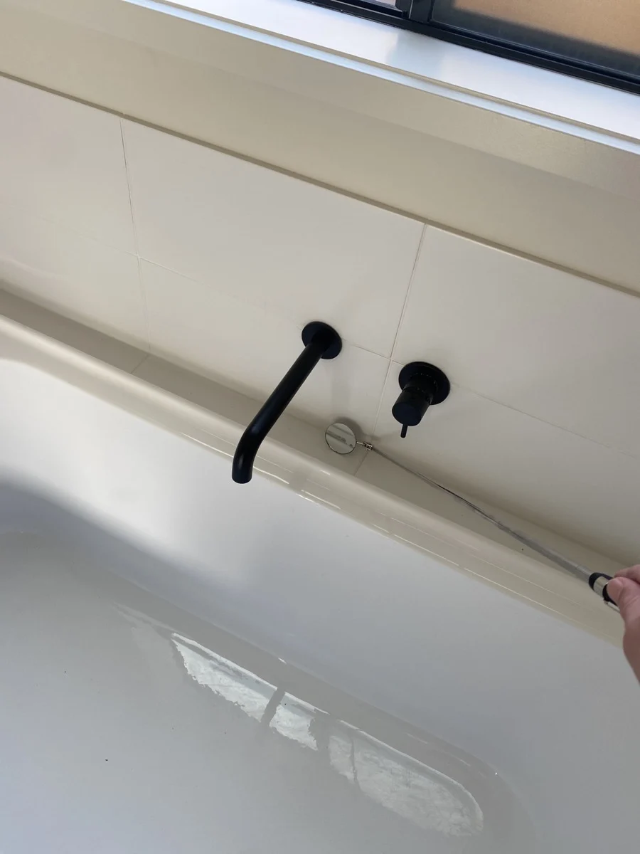 Hand holding a tape measure near black faucet and handle in a white bathtub. Sunlight from window creates a clean, minimalist look.