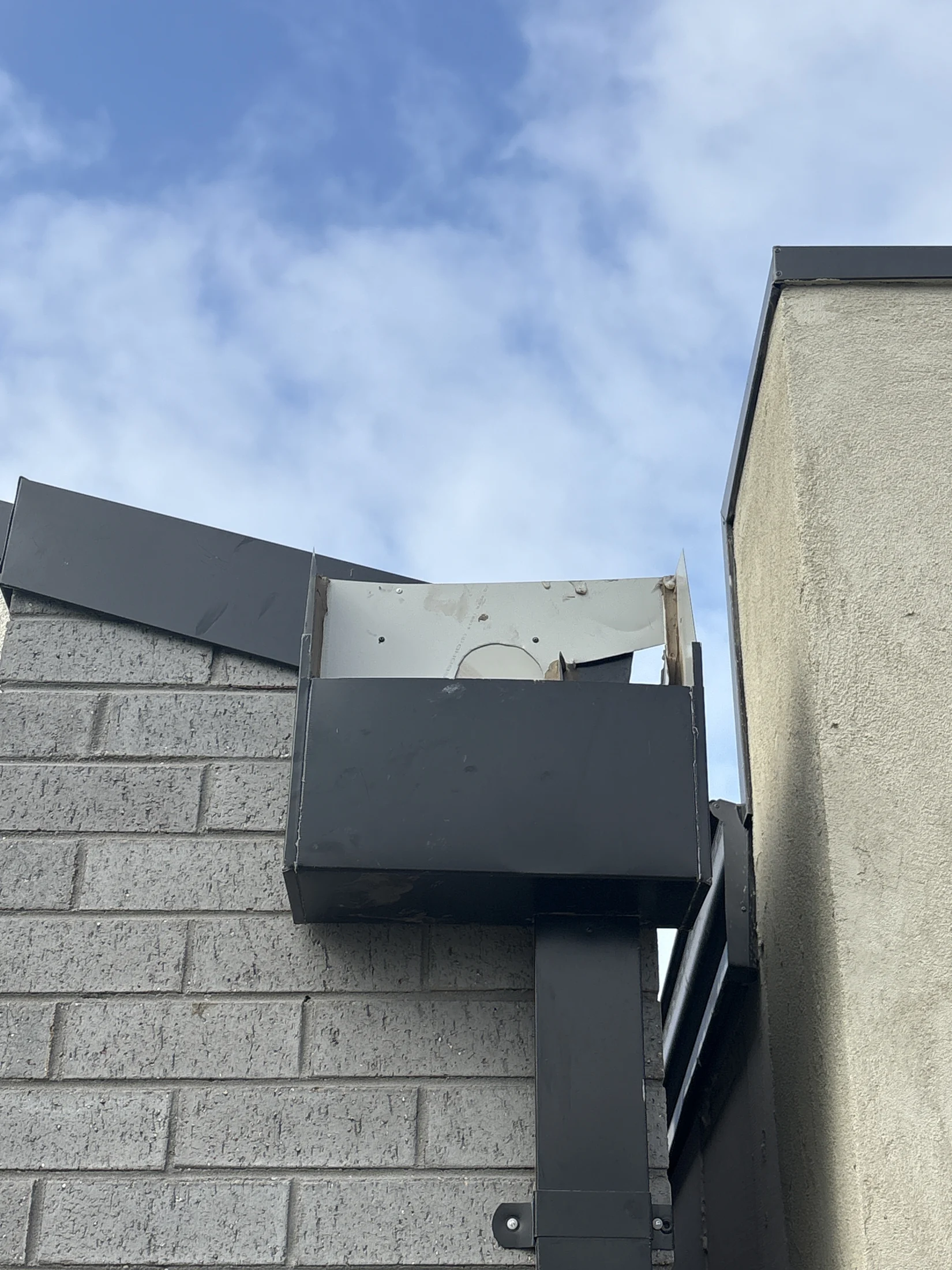 Rooftop corner with black ductwork and a damaged metal panel, against a brick wall and blue sky with clouds.