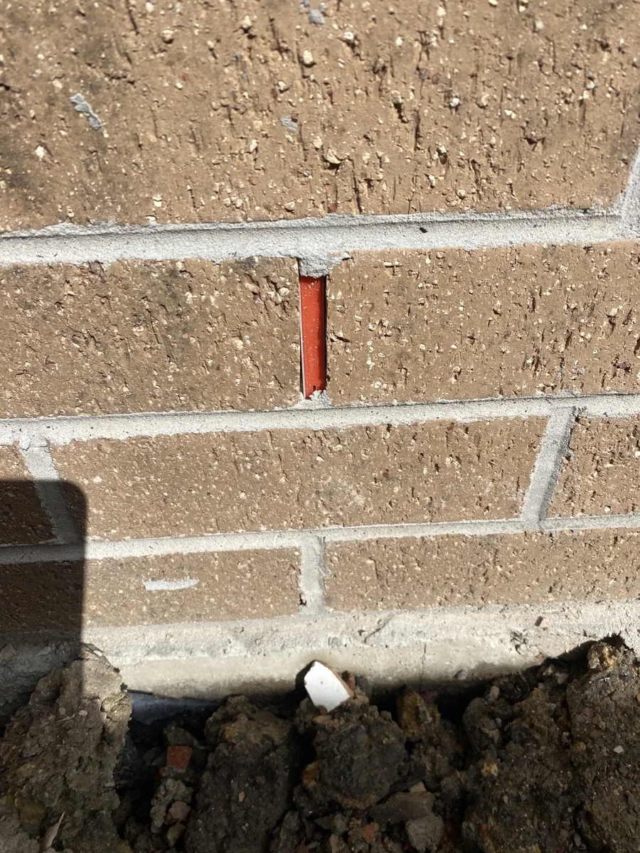 Close-up of a brick wall with a red pipe partially visible between bricks. Dirt and debris are at the base. Shadow casts on the wall.
