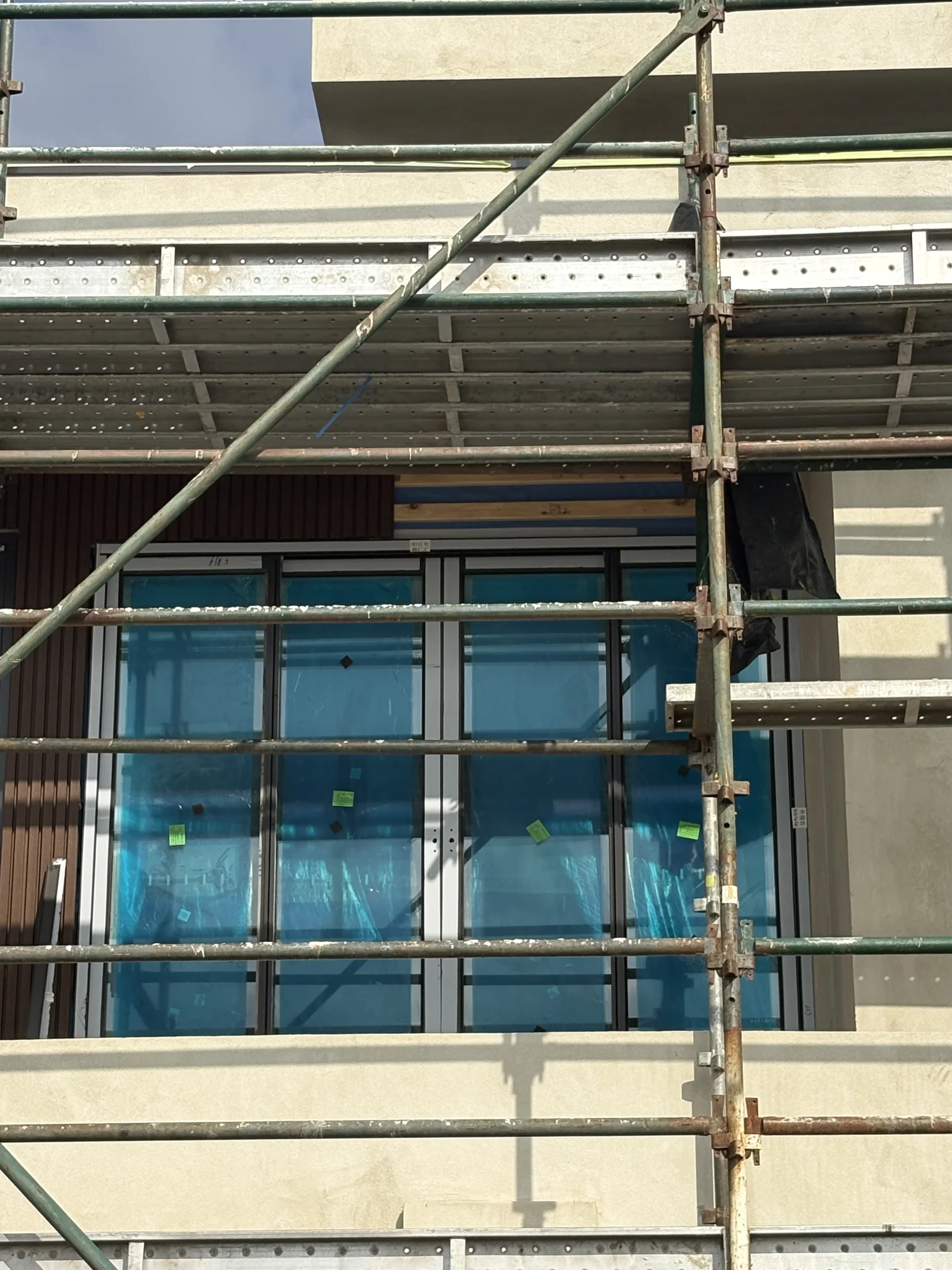 Scaffolding in front of building facade with teal-covered windows and beige walls. Clear sky above, construction setting visible.