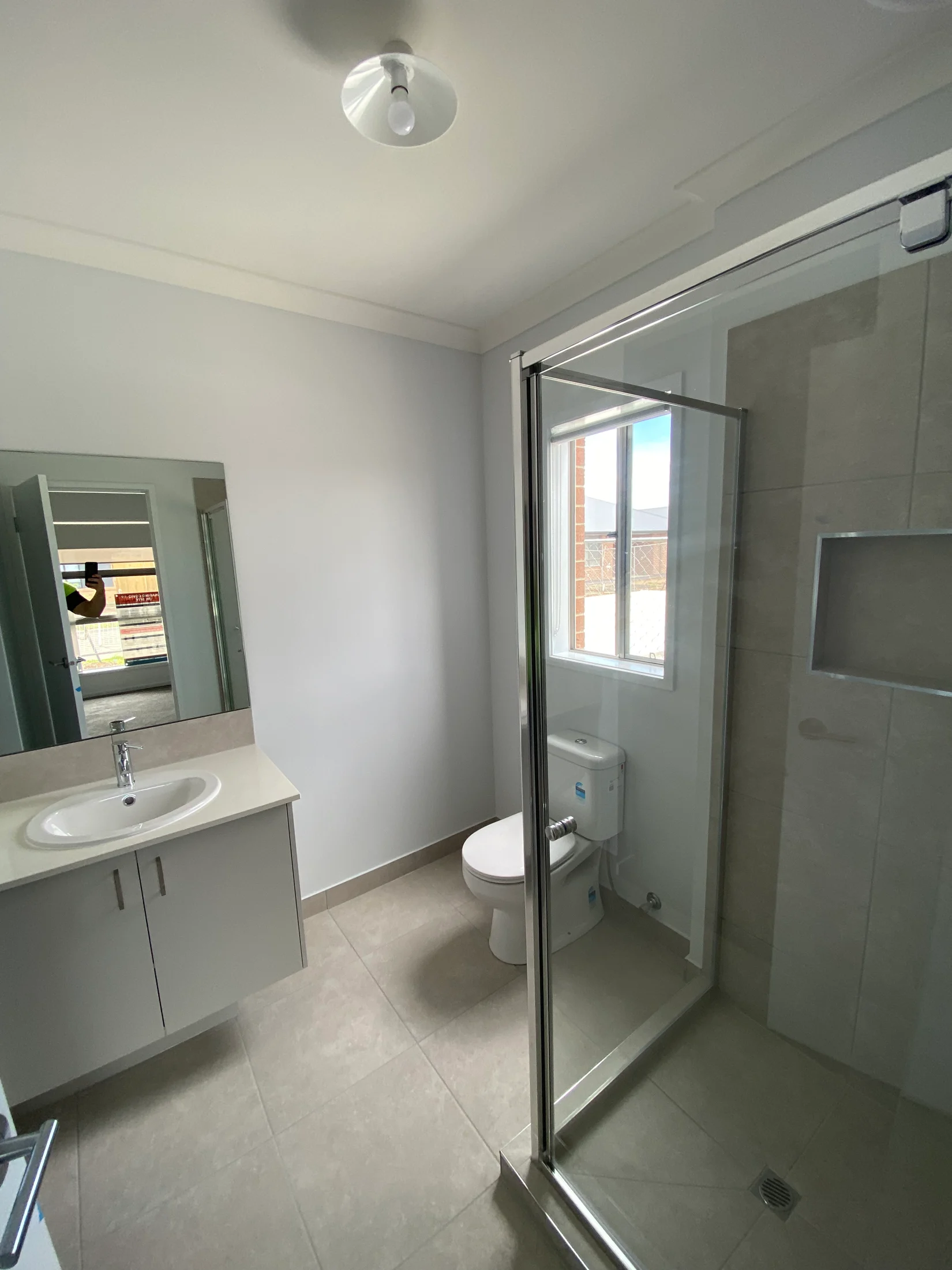 Modern bathroom with a shower enclosure, toilet, and sink. Neutral tiles and white walls create a clean, minimalist look. Natural light enters from a window.