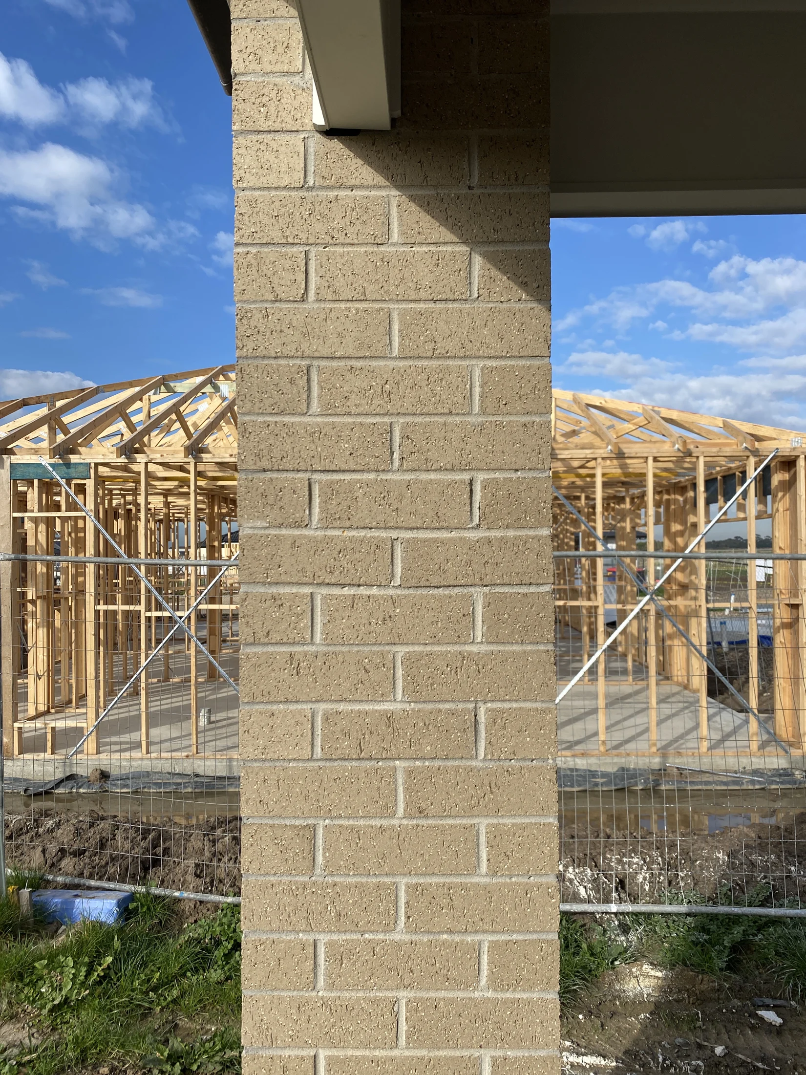 Brick column in foreground with wooden house frame construction in background. Blue sky with clouds overhead. Earth and grass visible below.
