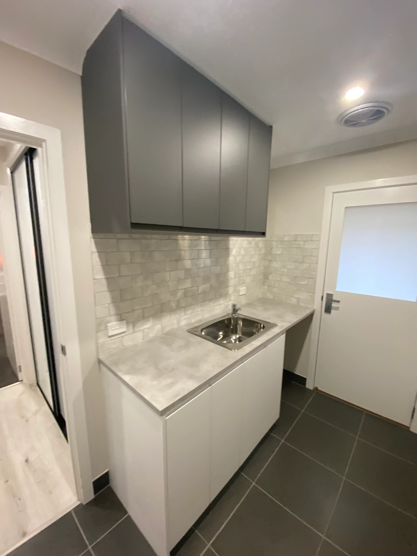 Modern laundry room with gray cabinets, white tile backsplash, and a stainless steel sink. Gray tiled floor and a white frosted door.
