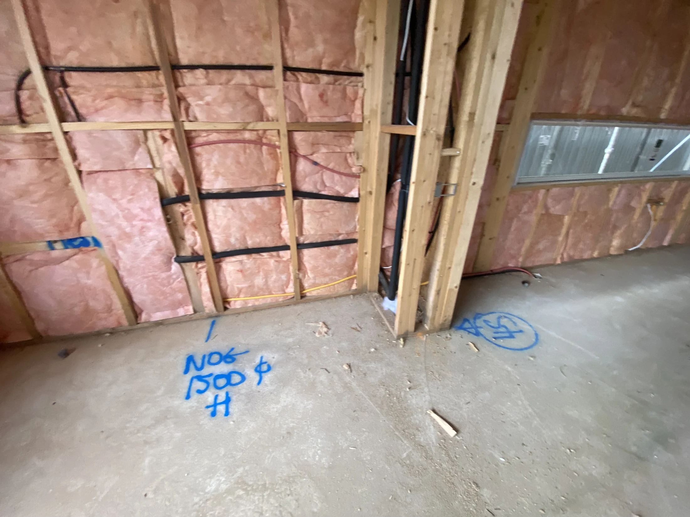 Exposed wooden framing with pink insulation in a construction site, blue markings on concrete floor, window on the right, unfinished look.