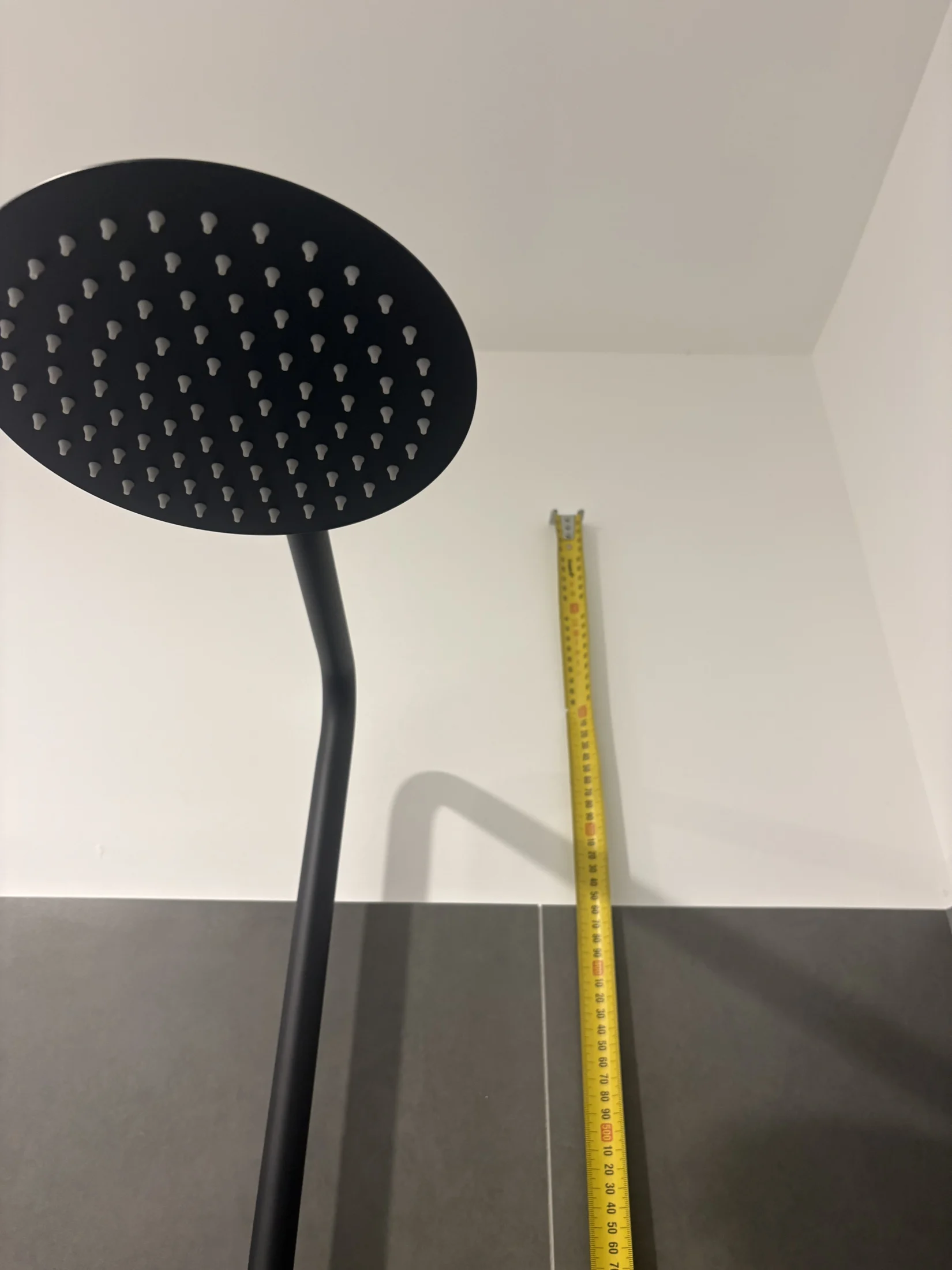 Black showerhead and yellow measuring tape against a white wall and dark tiles. The scene is minimalist with contrasting colors.