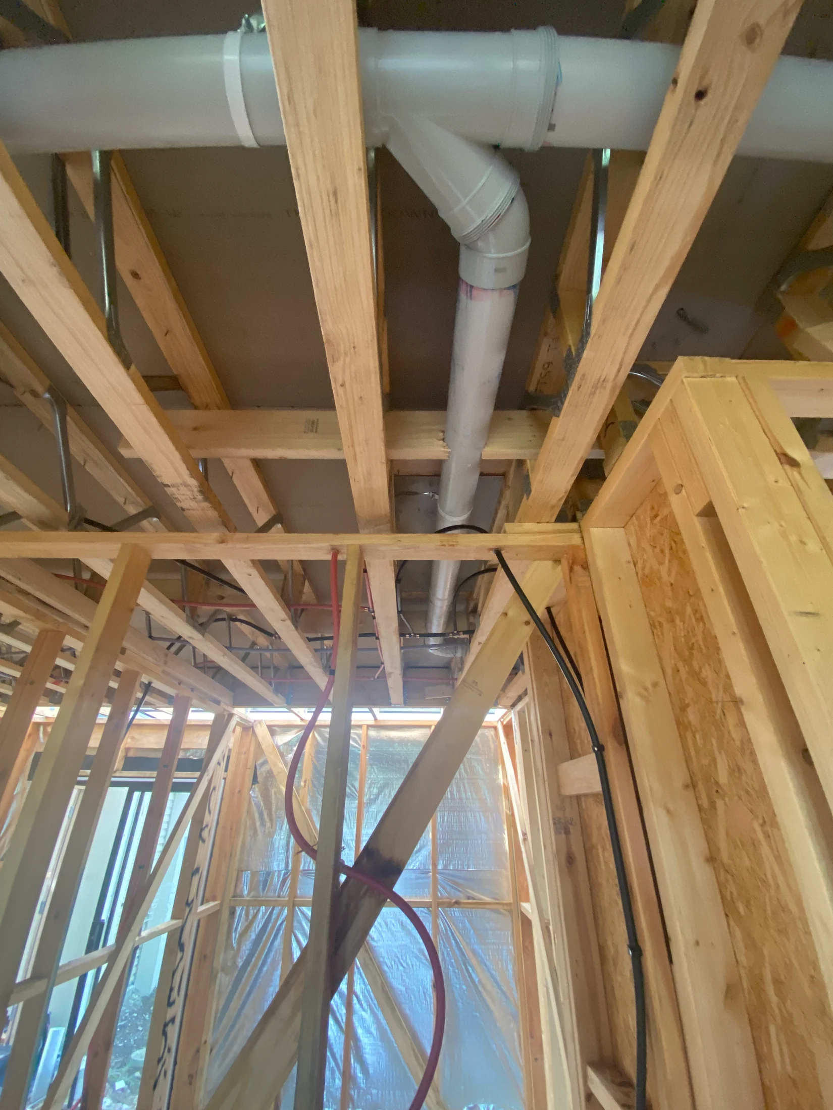 Wooden framing of a building under construction, with exposed pipes and wires. Sunlight filters through a plastic-covered window.