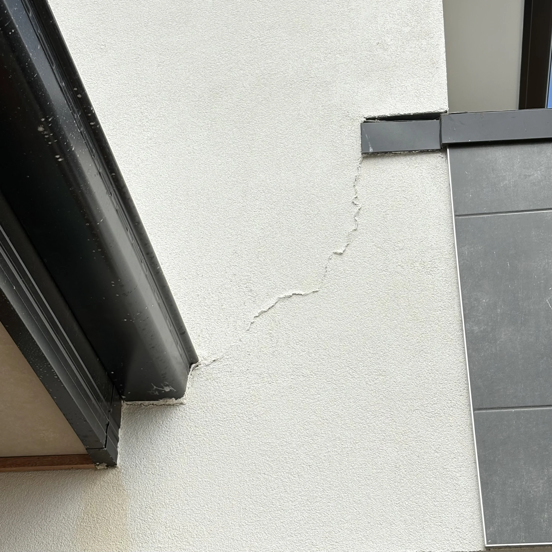 Close-up of a building corner showing a vertical crack in white stucco near a black gutter and gray panel. No text. Neutral mood.