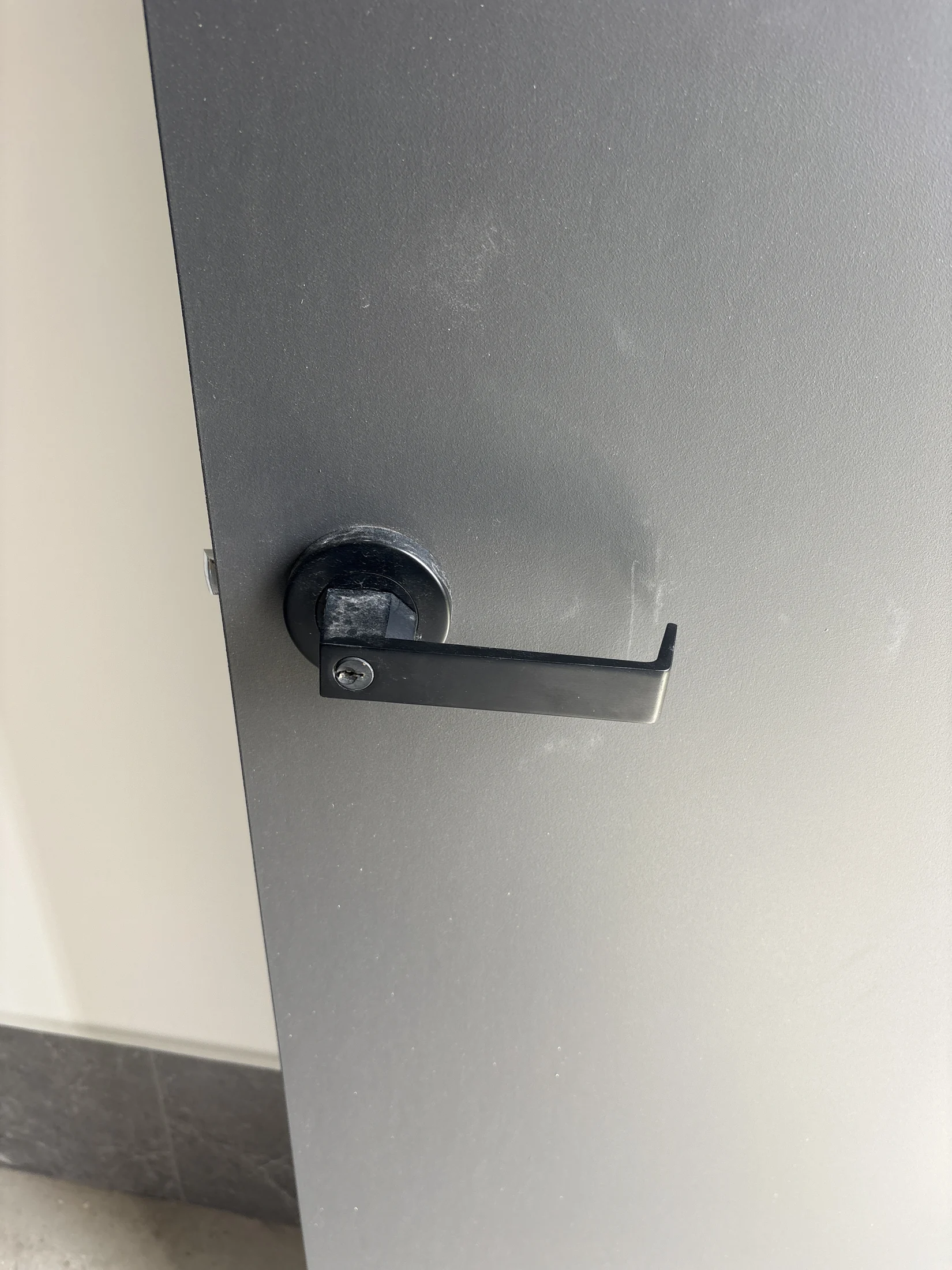 A black metal lever door handle on a gray door in a minimalistic indoor setting. Light scuffs are visible on the surface.