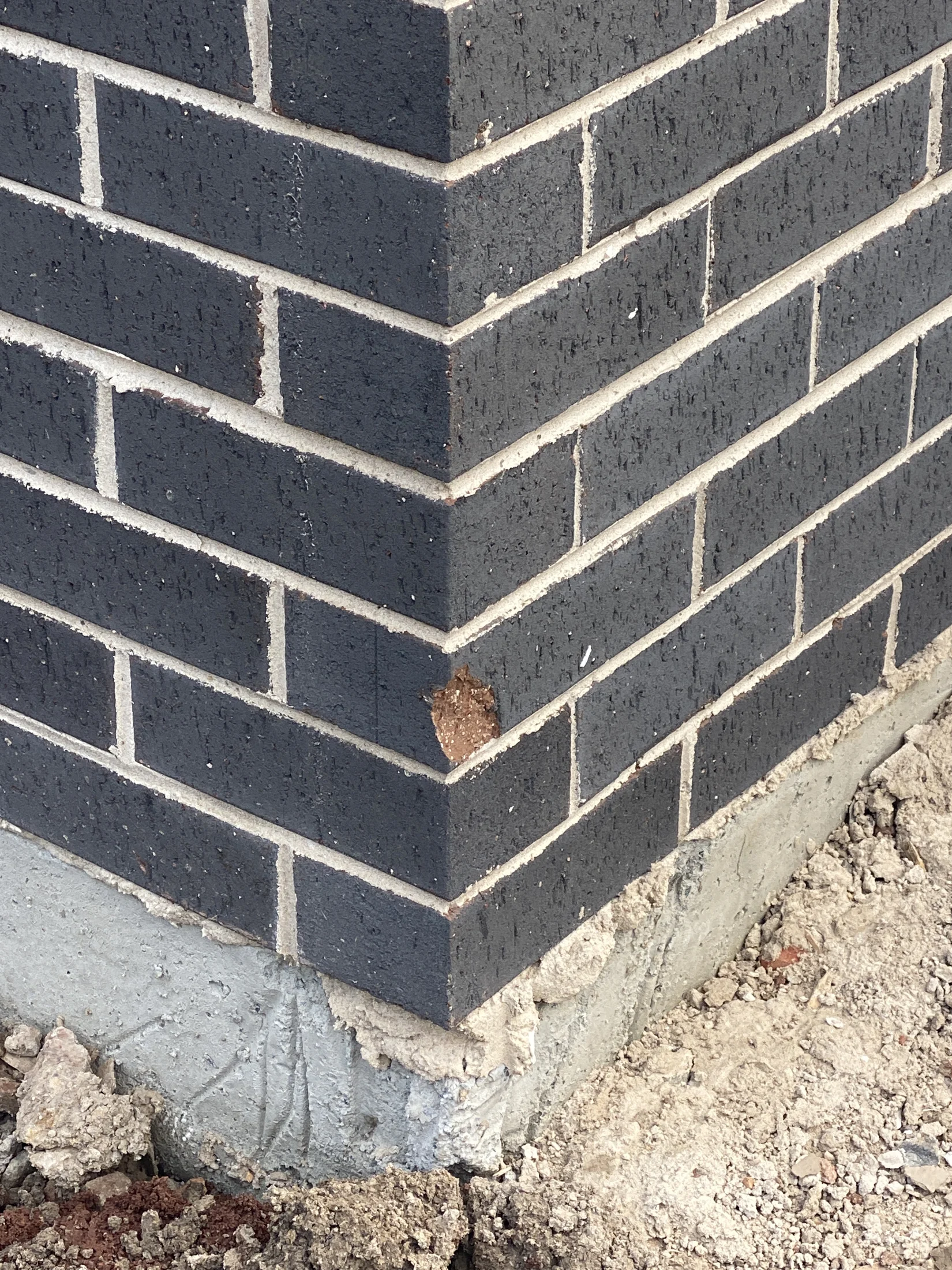 Corner of a brick wall with dark bricks and white mortar. A small chip is missing. Wall sits on a concrete foundation, surrounded by dirt.