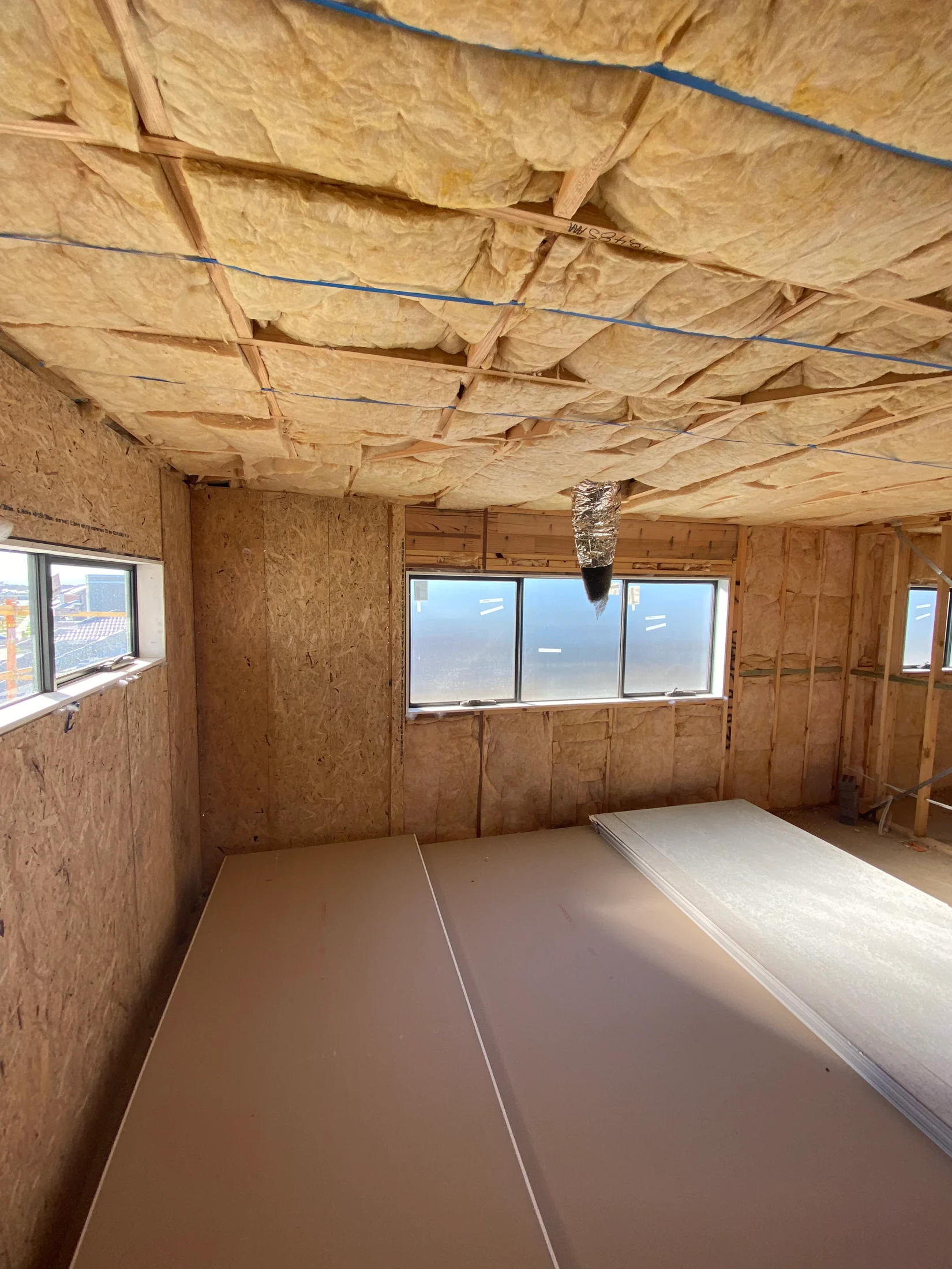 Construction room with yellow insulation on ceiling, wooden walls, and stacked drywall sheets. Sunlight through windows creates a bright mood.