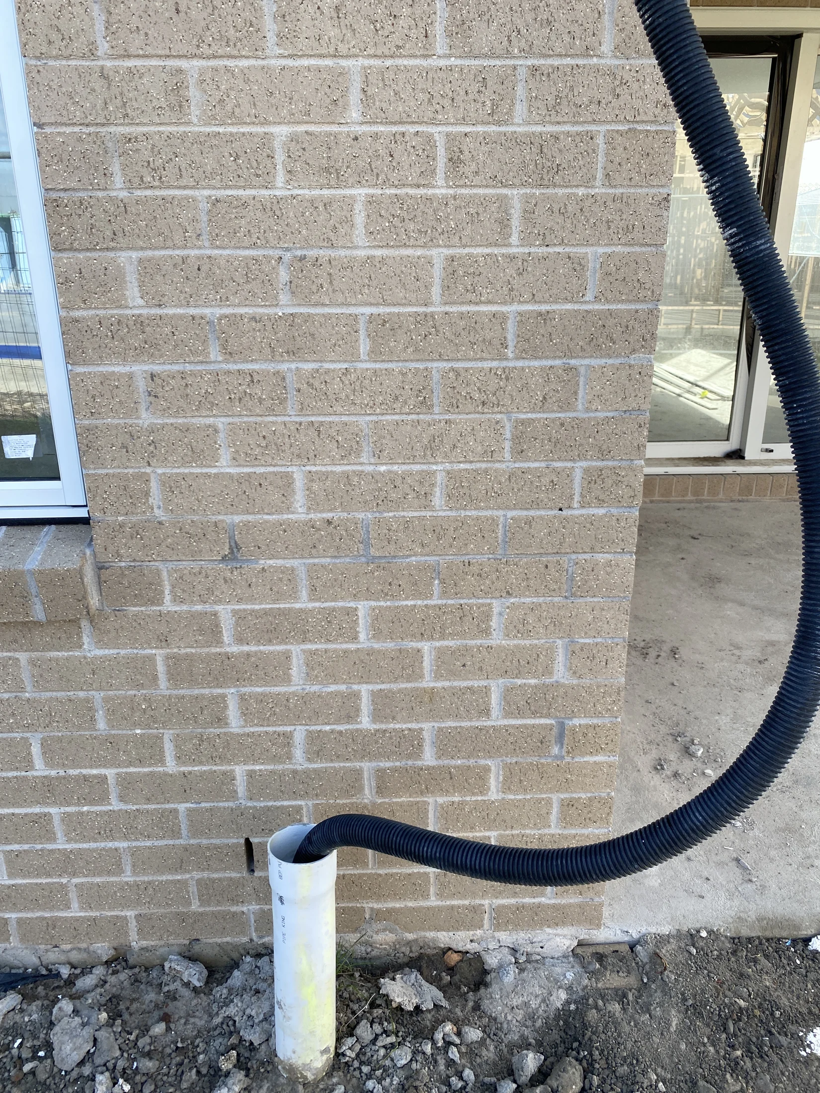 A black corrugated pipe enters a white PVC pipe against a tan brick wall. The ground is rough, and a sliding glass door is partially visible.