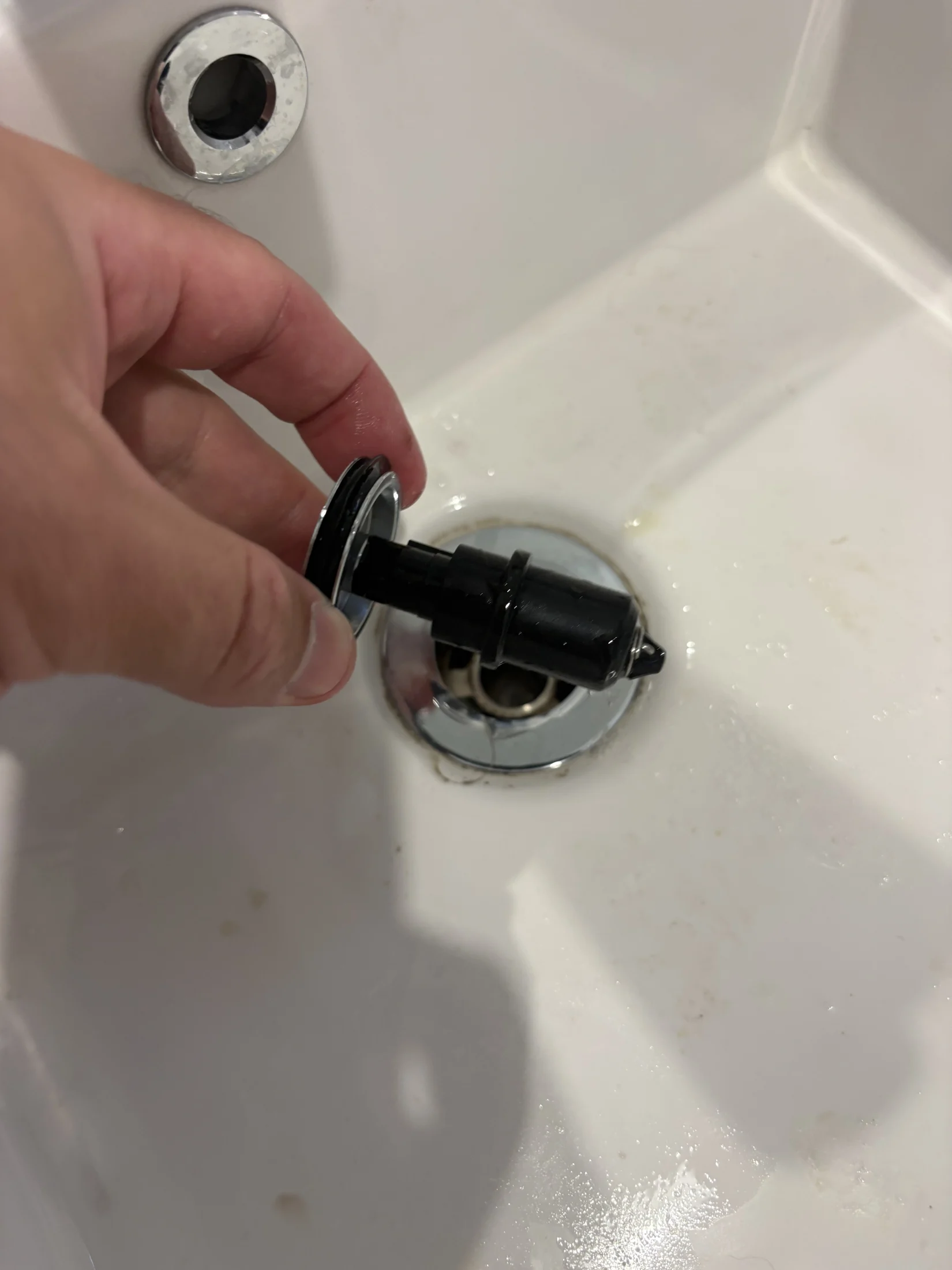 Hand holding a sink stopper over a white bathroom basin with water stains. Chrome drain visible in the background. Bright lighting.