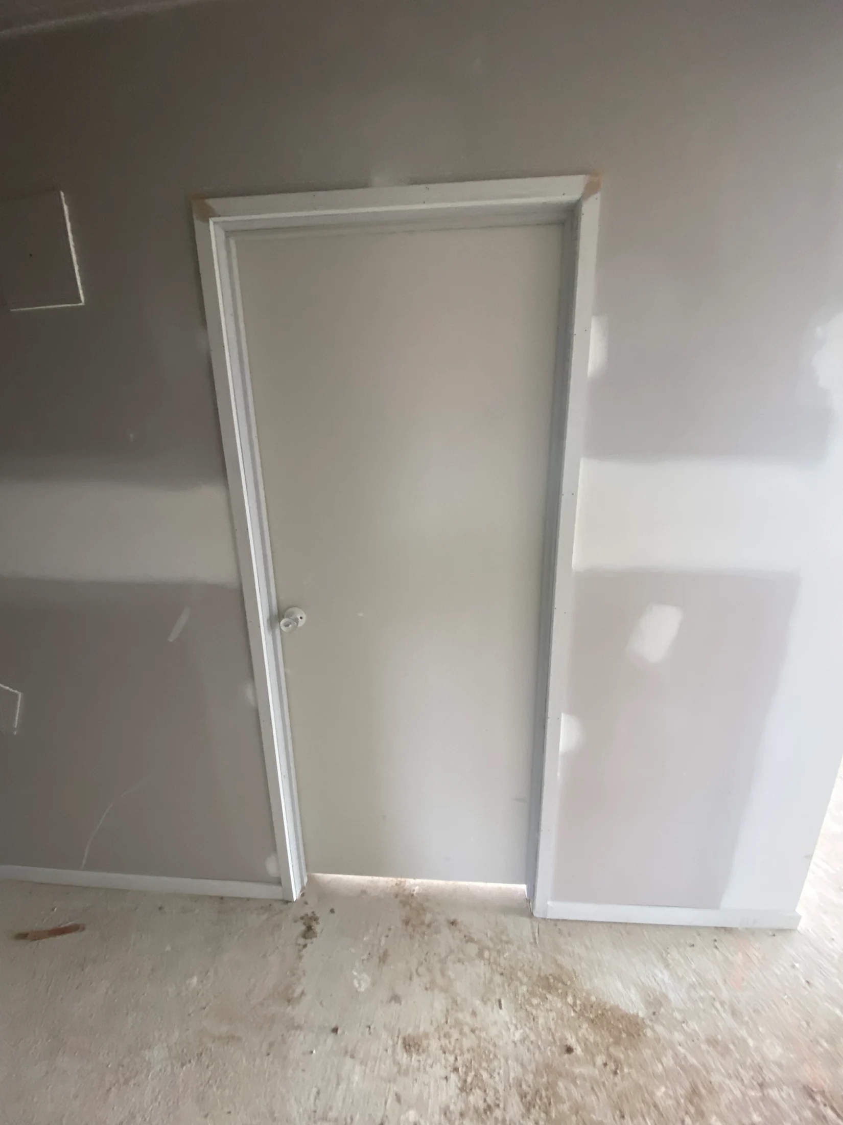 Plain white door in a partially finished room with drywall and a dusty concrete floor. No visible text or notable colors.