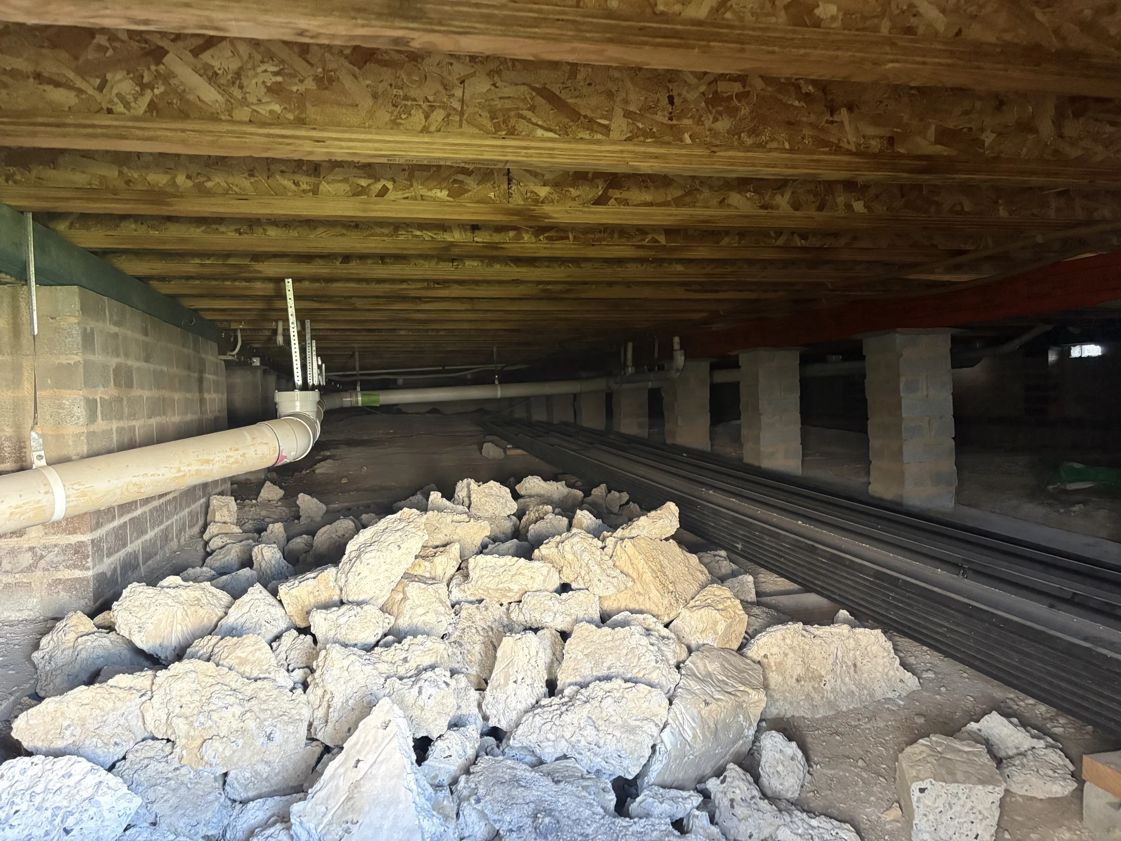 Pile of rubble in a dimly lit crawlspace with exposed wooden beams and pipes. Concrete blocks support the structure. No visible text.