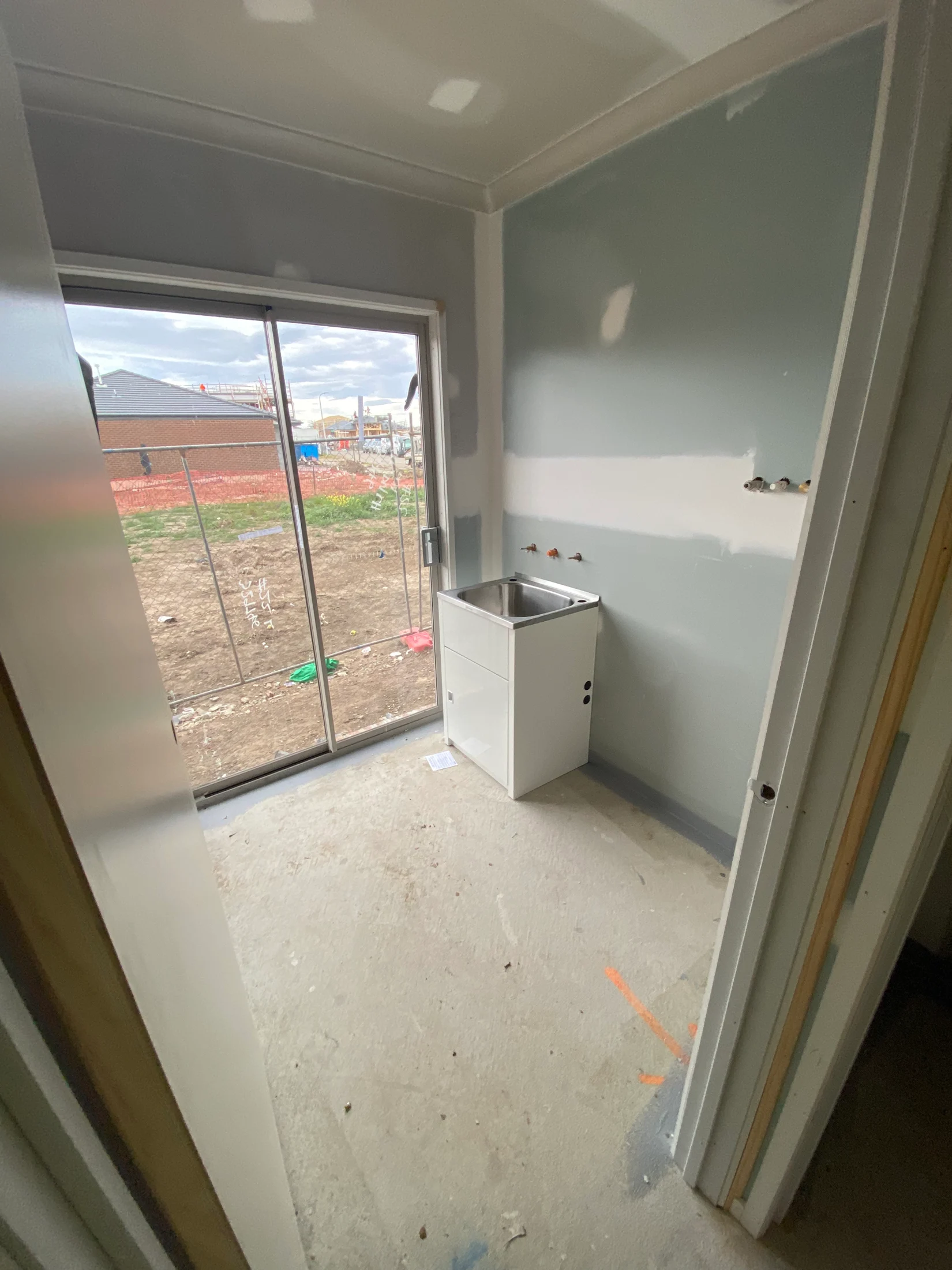 Small unfinished room with gray walls, a metal sink, and sliding glass door. Outside, a construction site is visible under a cloudy sky.
