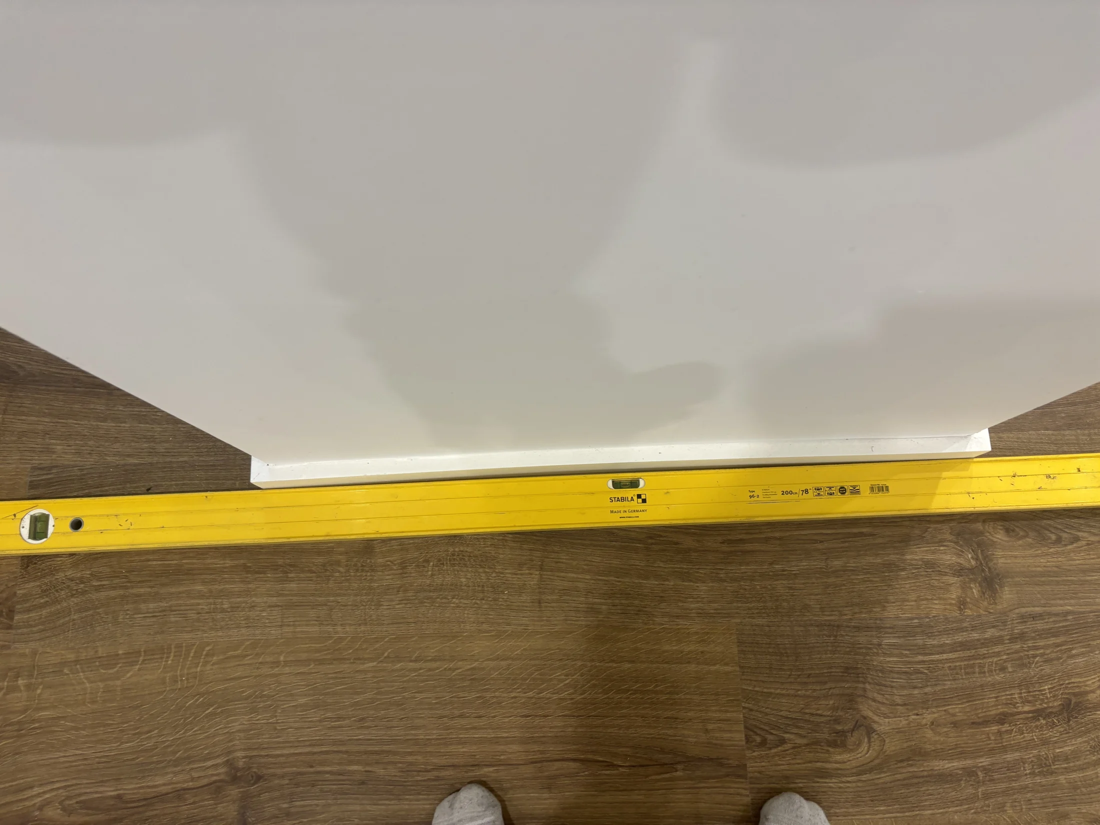 Yellow measuring level on wooden floor, leaning against a white wall. Visible text reads "Stabila." One foot in a sock is partially visible.