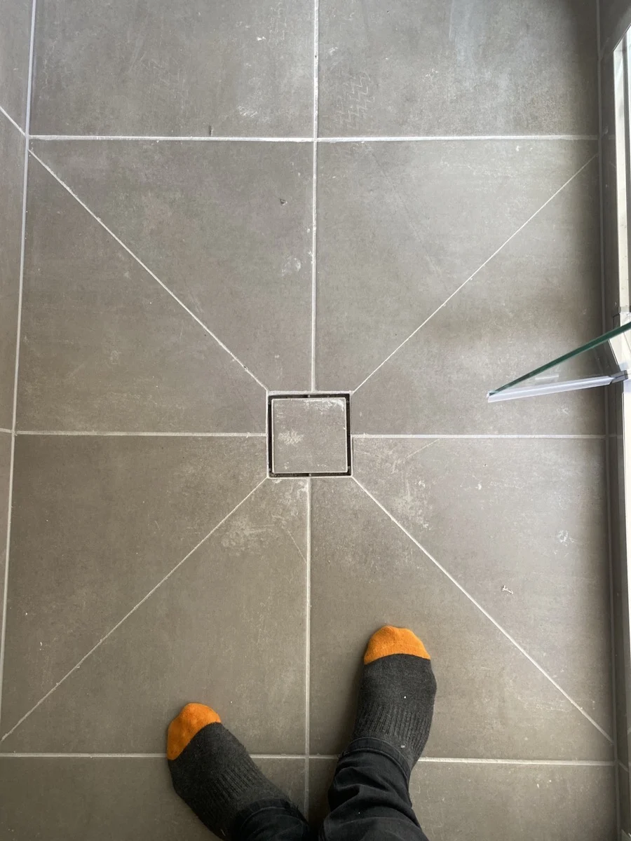 Feet in black socks with orange toes stand on gray tiled floor featuring intersecting white grout lines and a square tile in the center.