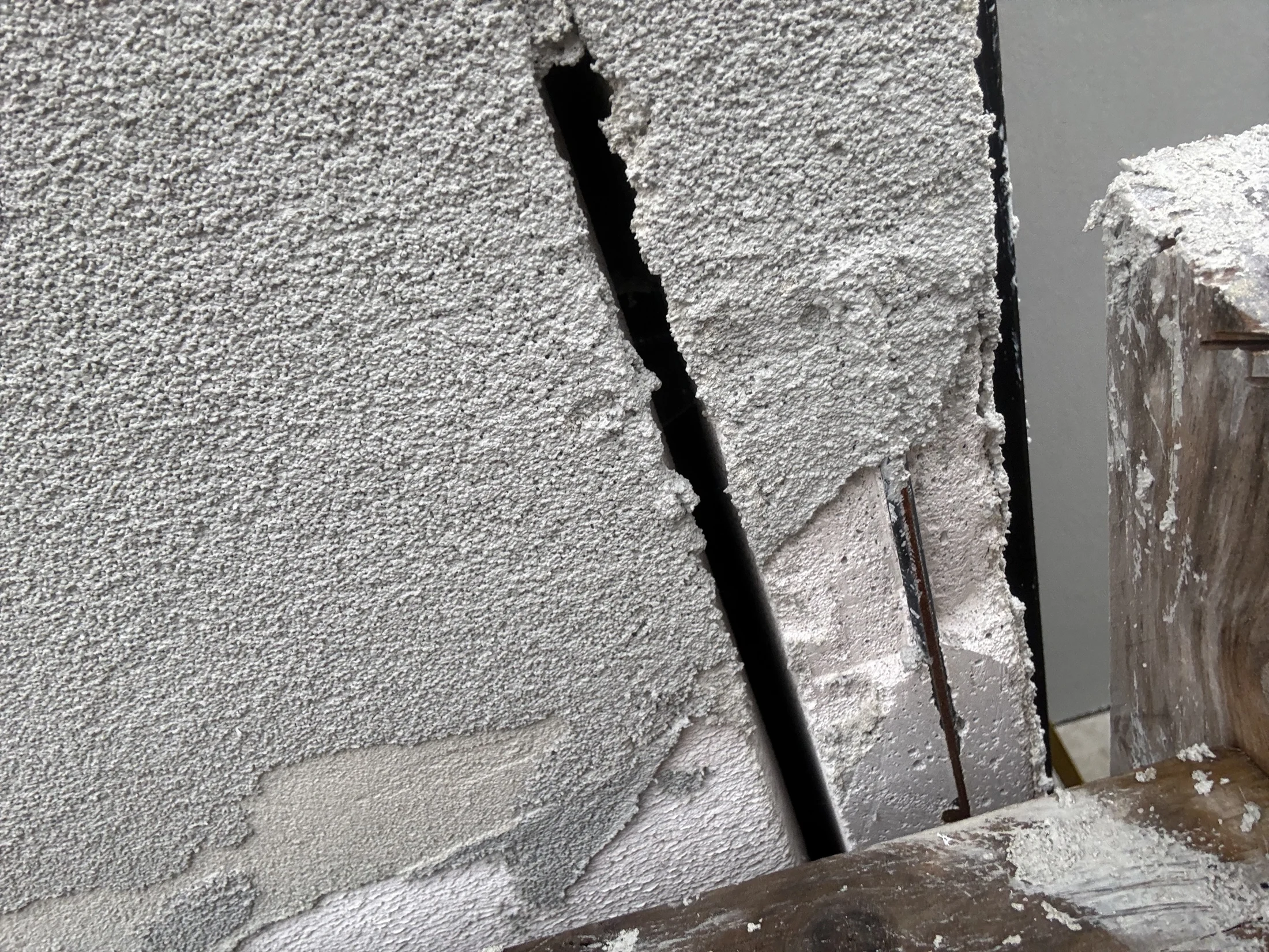 Close-up of a wall with a large crack, showing exposed reinforcement bars. The surface appears rough and weathered, with visible debris.