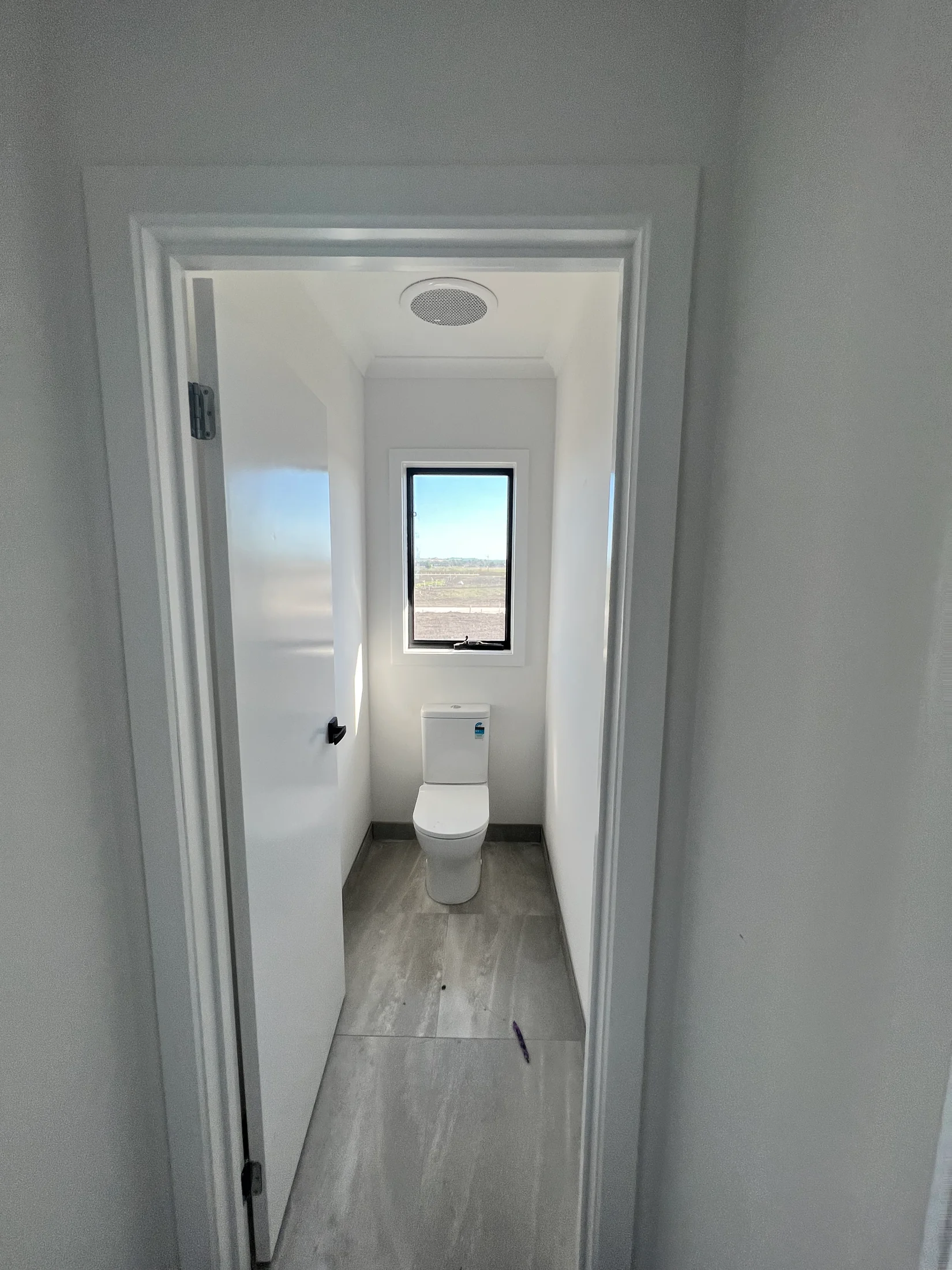 White bathroom with a toilet facing a window. Grey floor tiles and white walls create a clean, minimalist look. Bright natural light.
