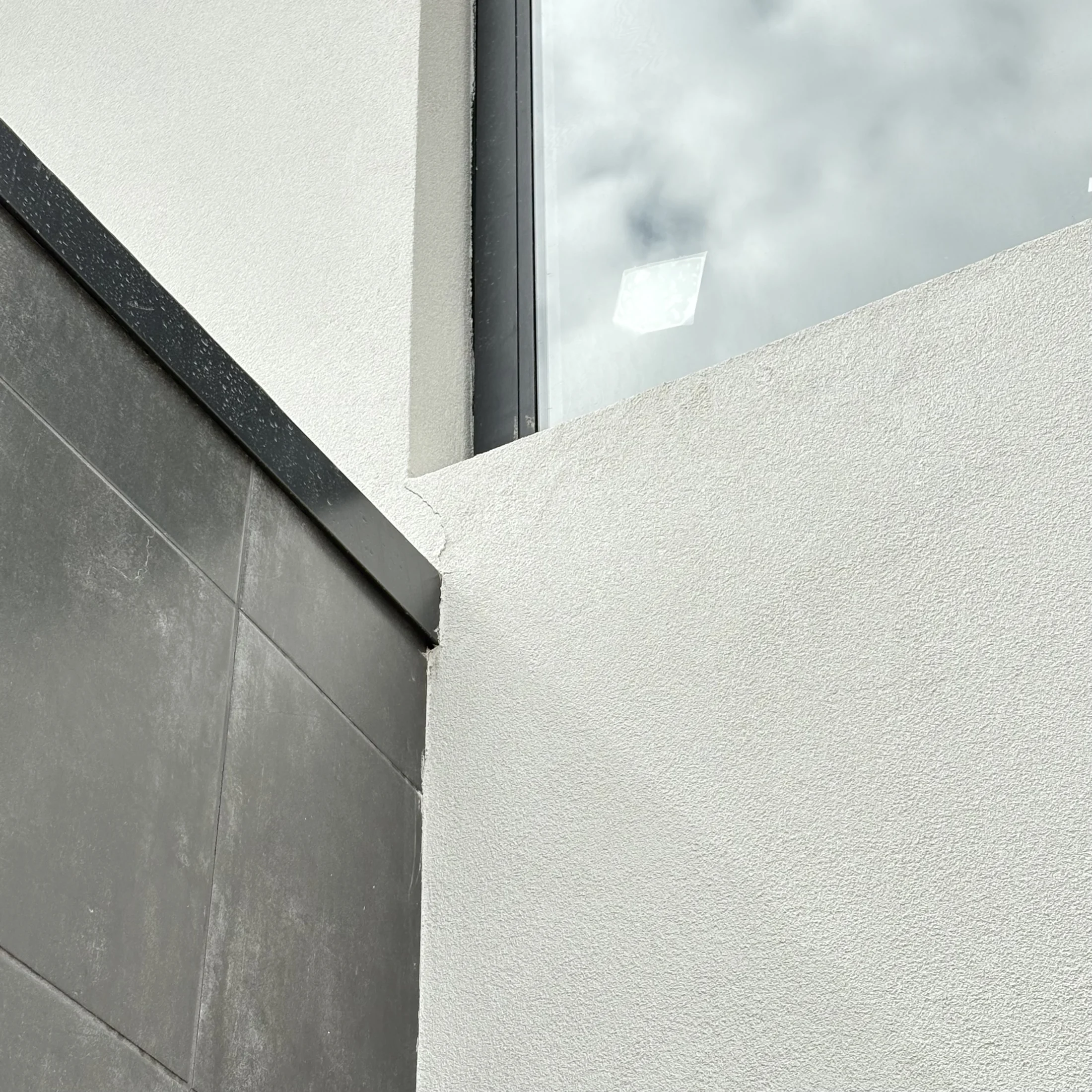 Corner of a modern building with textured white walls, gray tiles, and a large window reflecting a cloudy sky. Minimalist architectural design.