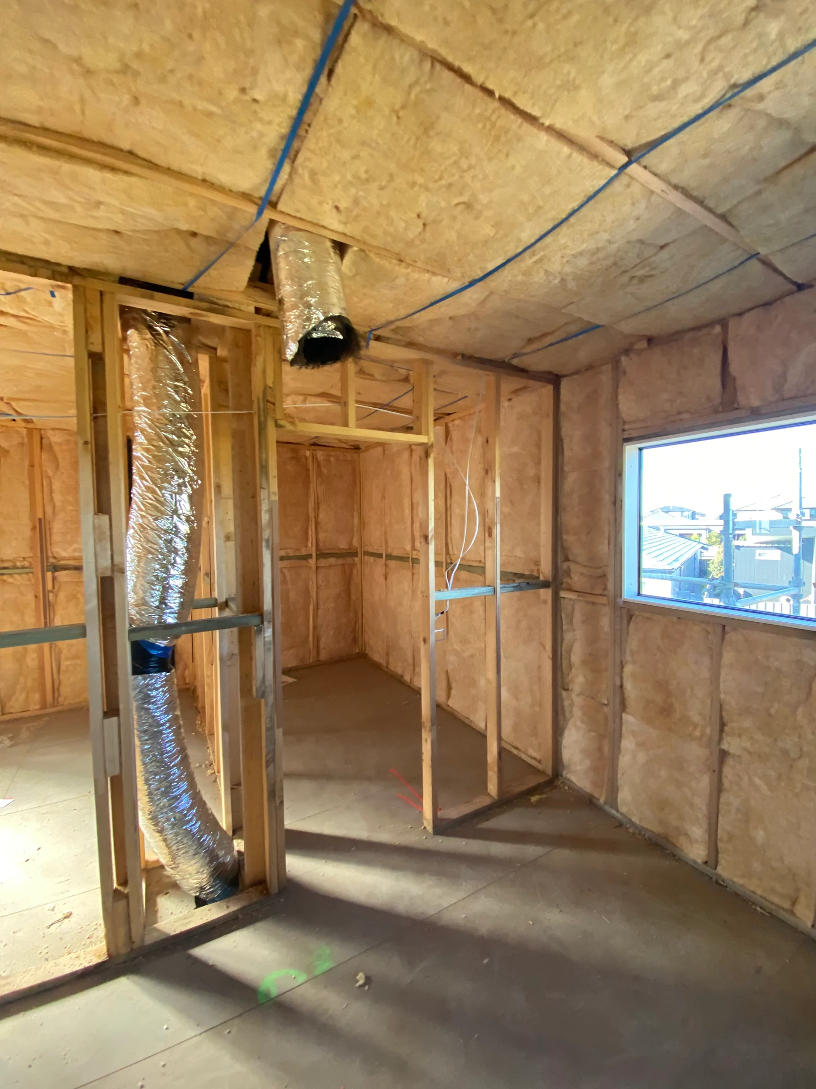 Unfinished room with wooden framing and insulation on walls and ceiling. A silver duct is visible. Natural light enters through a window.