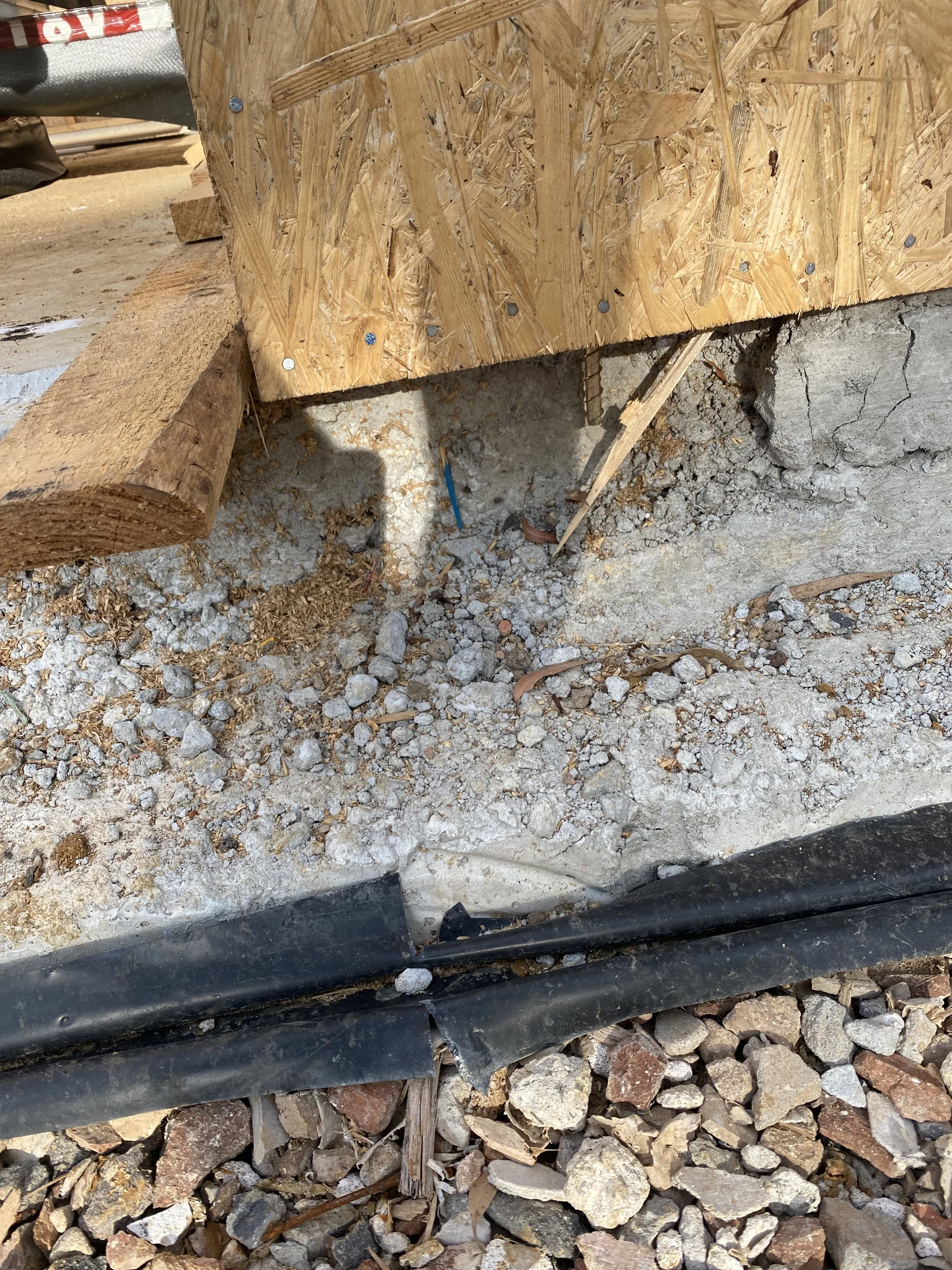 OSB board and wood beam on rocky ground with cracked concrete. Black tarp and scattered gravel visible in sunny outdoor setting.