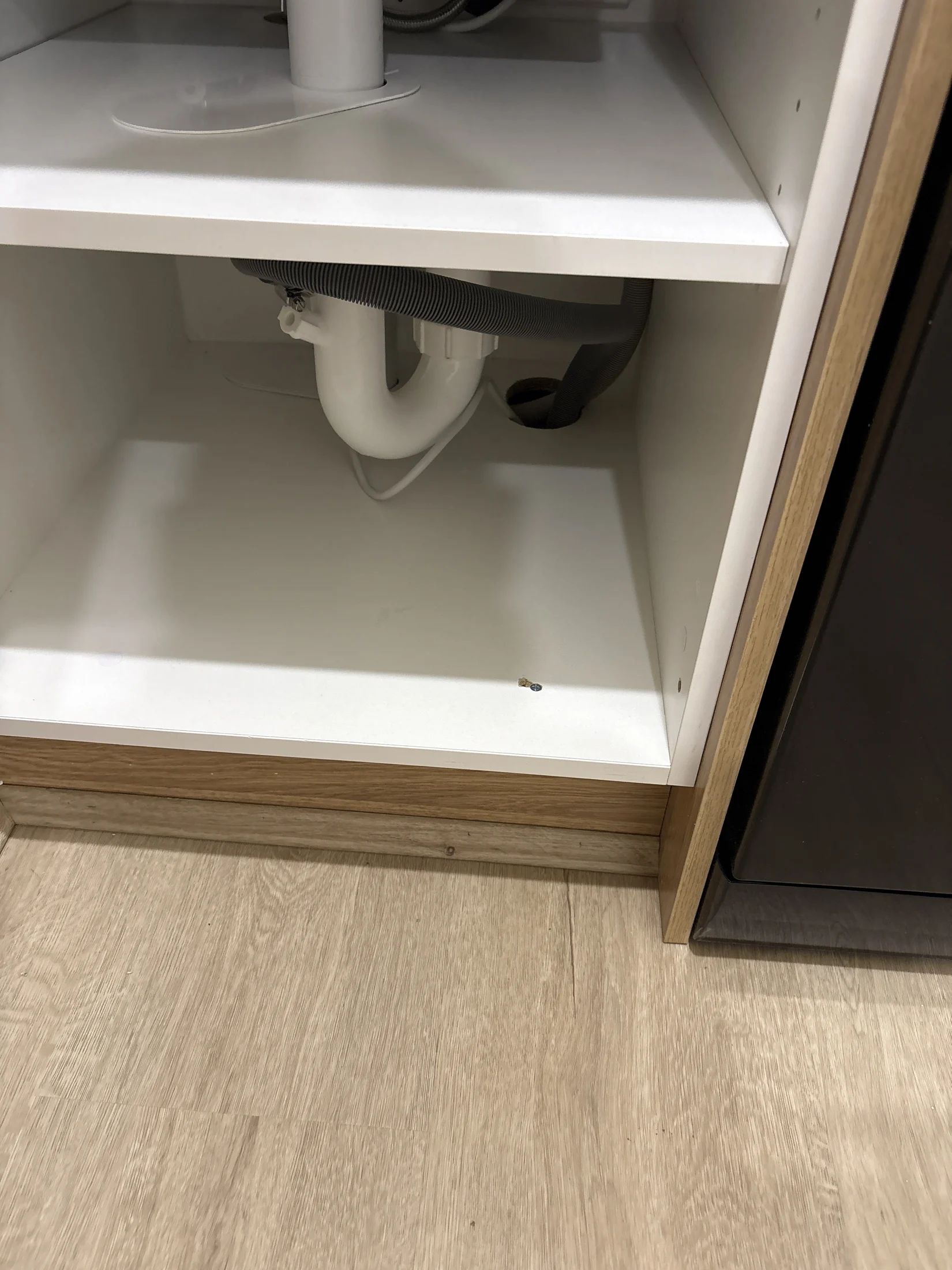 Under-sink cabinet with white pipes and a gray hose, wood floor visible. Bright lighting, minimal clutter, modern and clean setting.